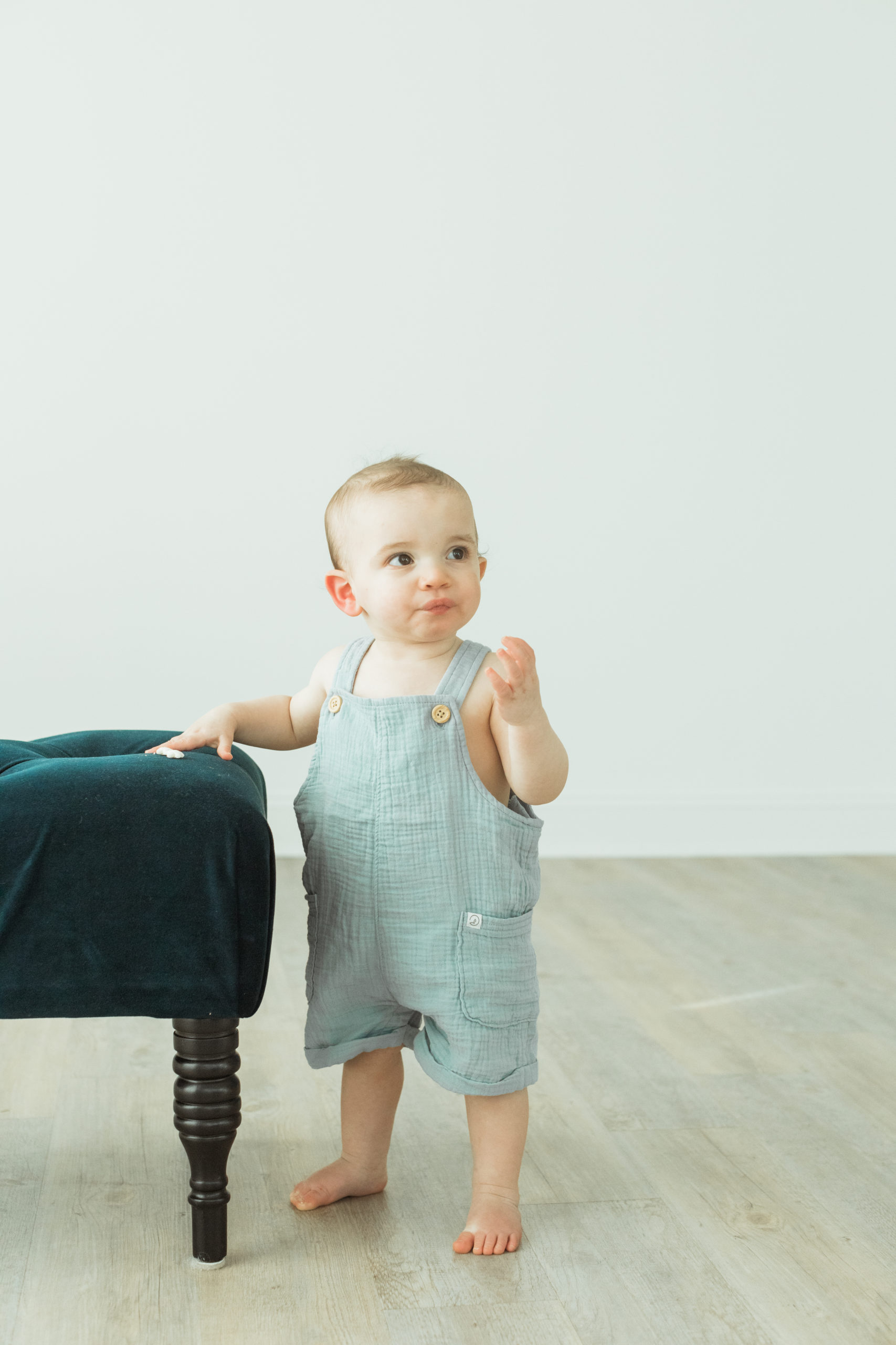 Photo of 1 year old baby boy standing near an emerald green velvet chair. Little boy's hand is resting in the chair while he's looking away from camera to the side. Baby boy barefoot in baby blue linen sleeveless overalls. Cute baby smile.