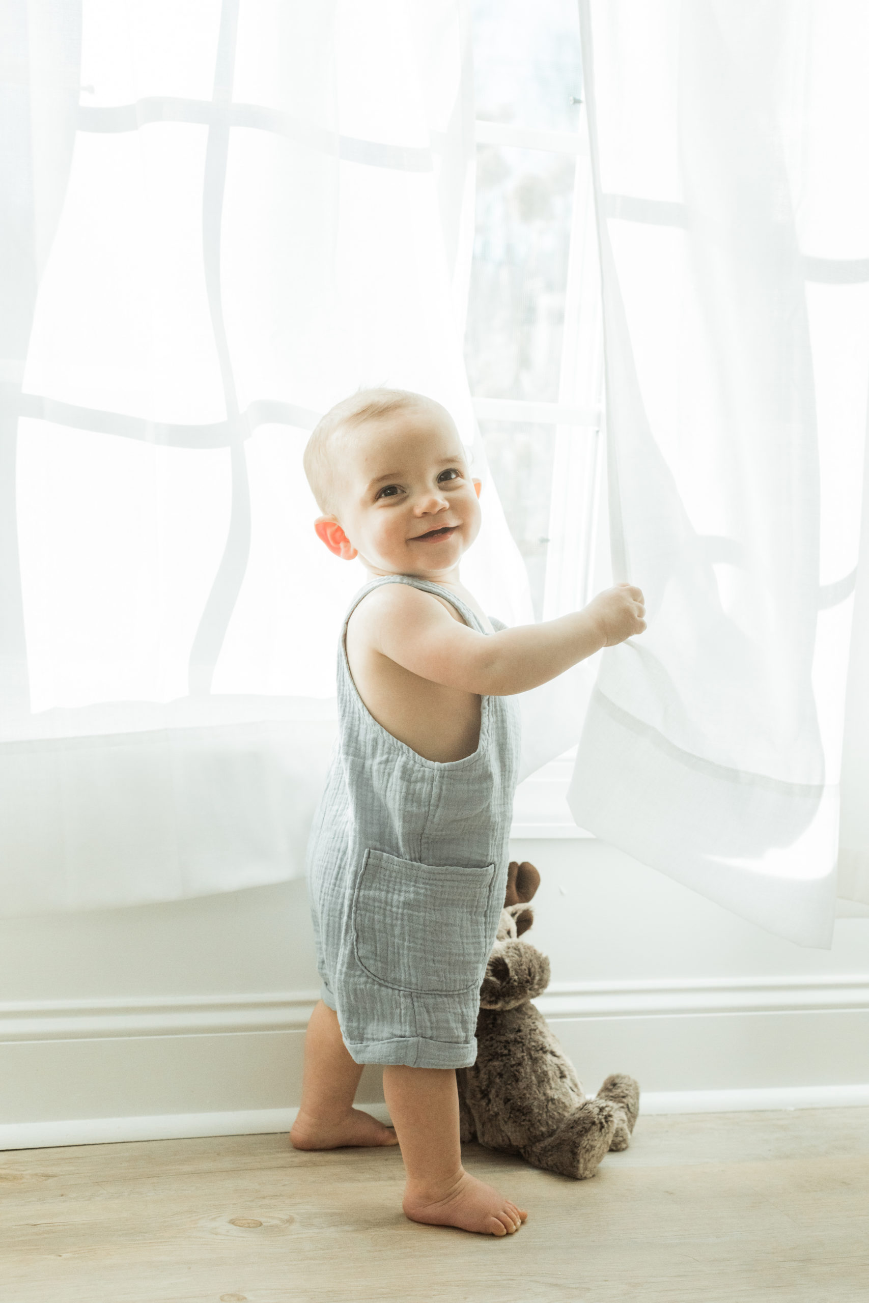 Photo of 1 year old baby boy standing near a window looking at the camera while playing with white curtains. Stuffed moose animal laying on the floor near his feet. Baby boy barefoot in baby blue linen sleeveless overalls. Cute baby smile.