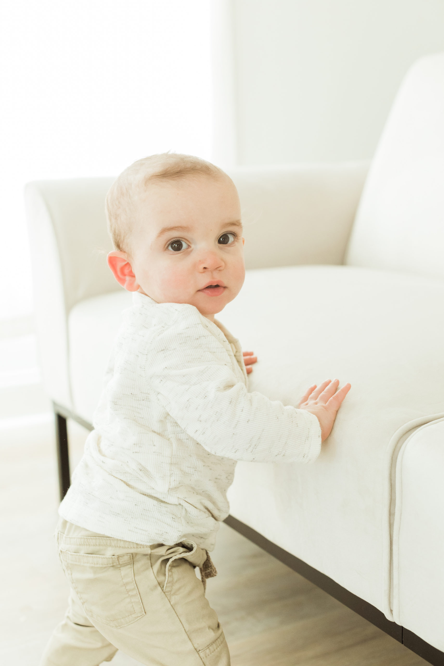 One year old baby boy standing up and leaning on light cream sofa chair. Little boy wearing light cream long sleeve and khaki pants. Photographed in studio under natural light.

