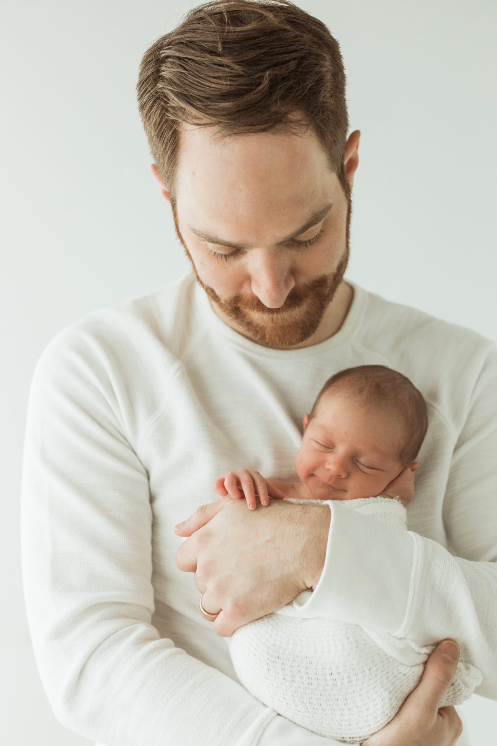 Dad in white long sleeve sweater holding his newborn baby girl wrapped up in white blanket near his chest. Both dad and baby facing the camera. Dad looking down at his daughter. Newborn baby girl with eyes closed smiling and hand on dad's.