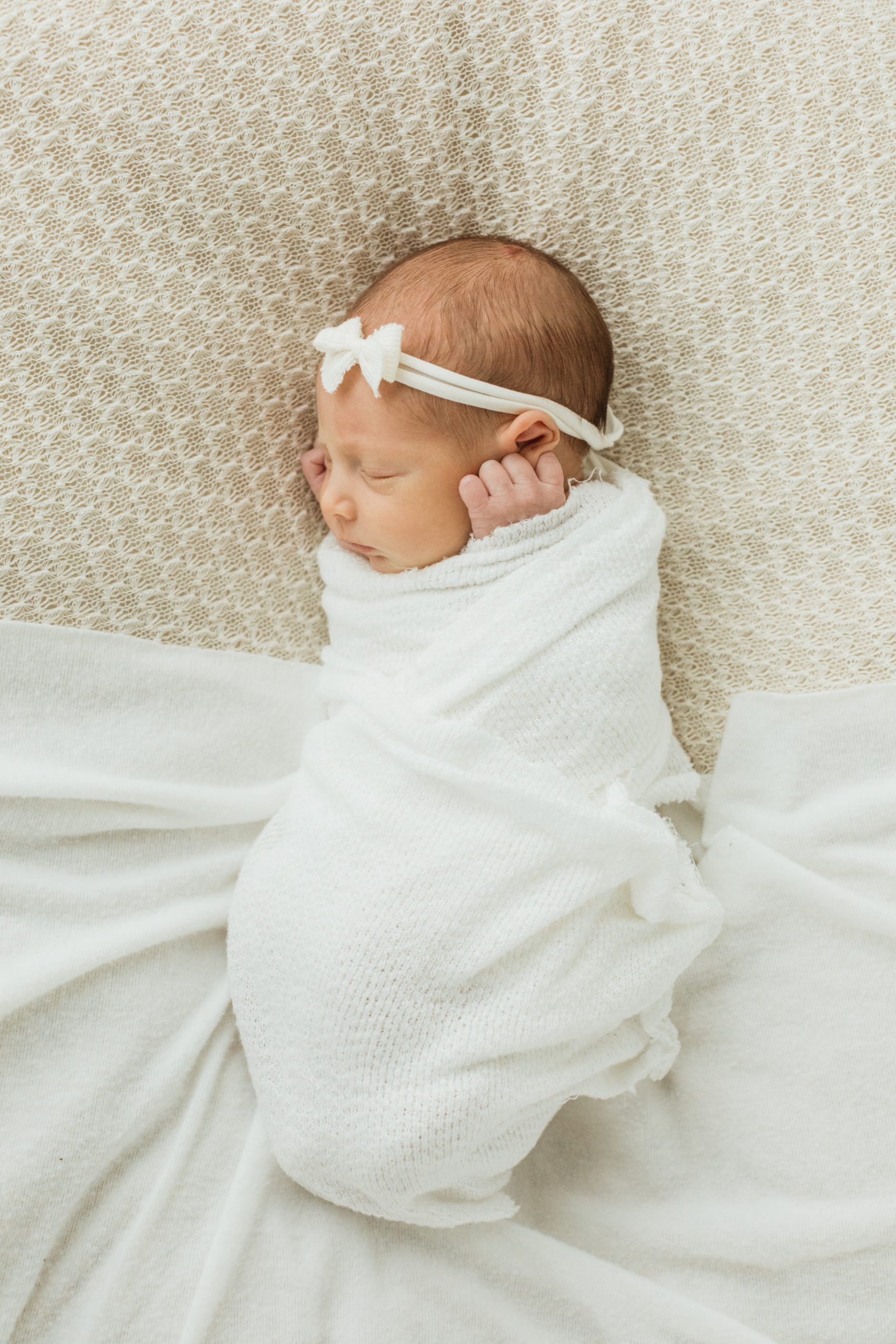 Sleepy newborn baby girl wrapped in white blanket in white bed with white headband.