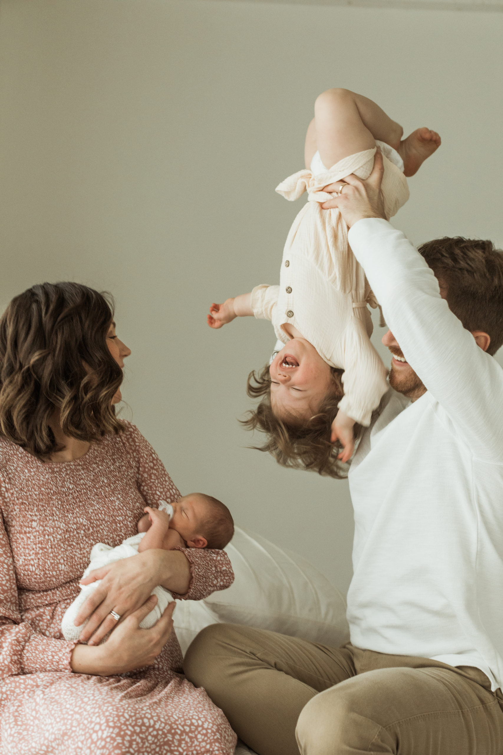 Photo of family of four sitting down on bed for their newborn photo session. Mama with loose curls wearing a pink and white printed long sleeve dress. Mama holding on to newborn baby. Dad wearing white long sleeve seater with khaki pants holding his oldest daughter up in the air upside-down . Little girl in white dress laughing.