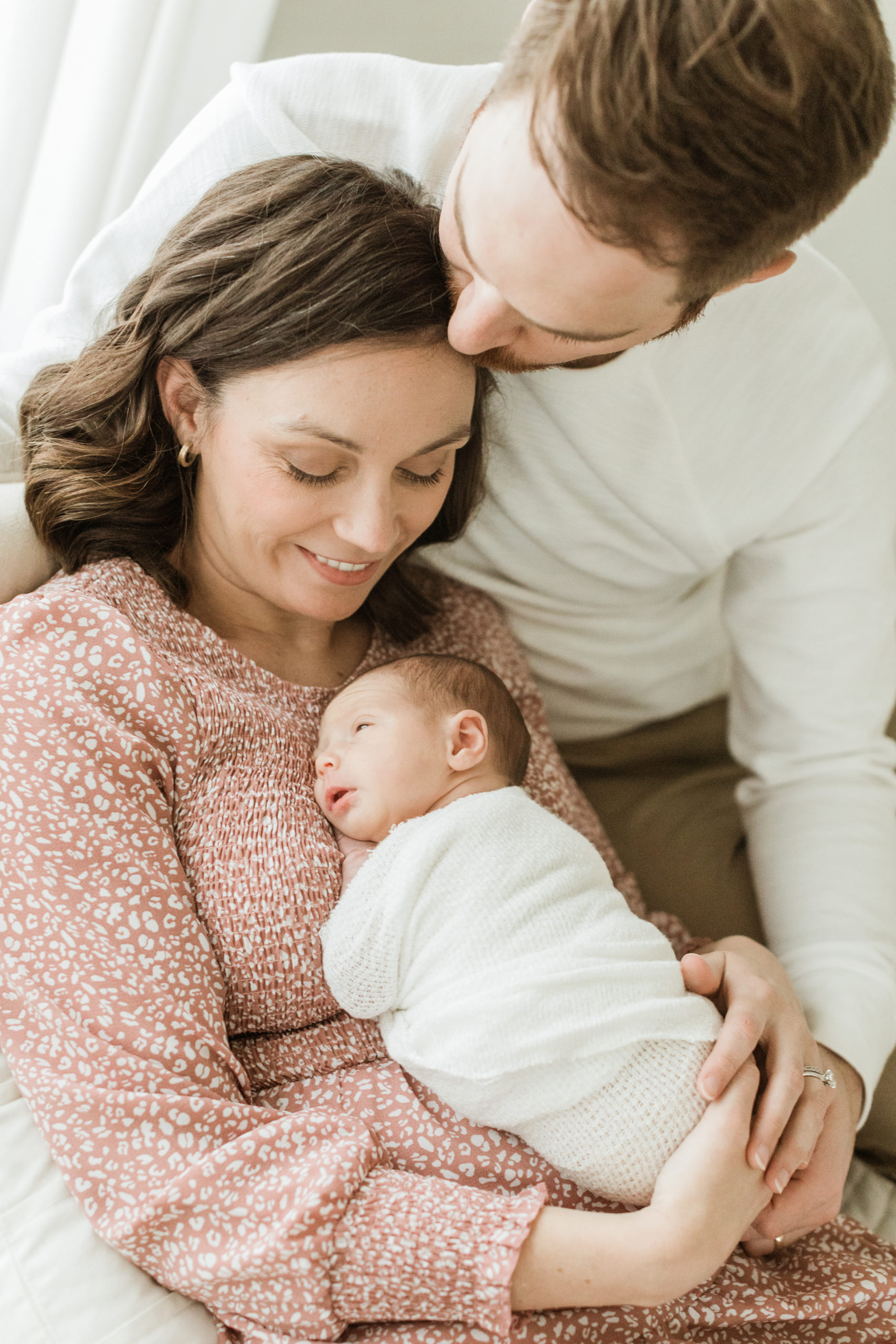 Mama and dad with their newborn daughter.mama in pink and white leopard printed dress holding her baby girl wrapped in white blanket on her chest. Dad wearing cream long sleeve and khaki pants. Dad kissing wife's forehead.