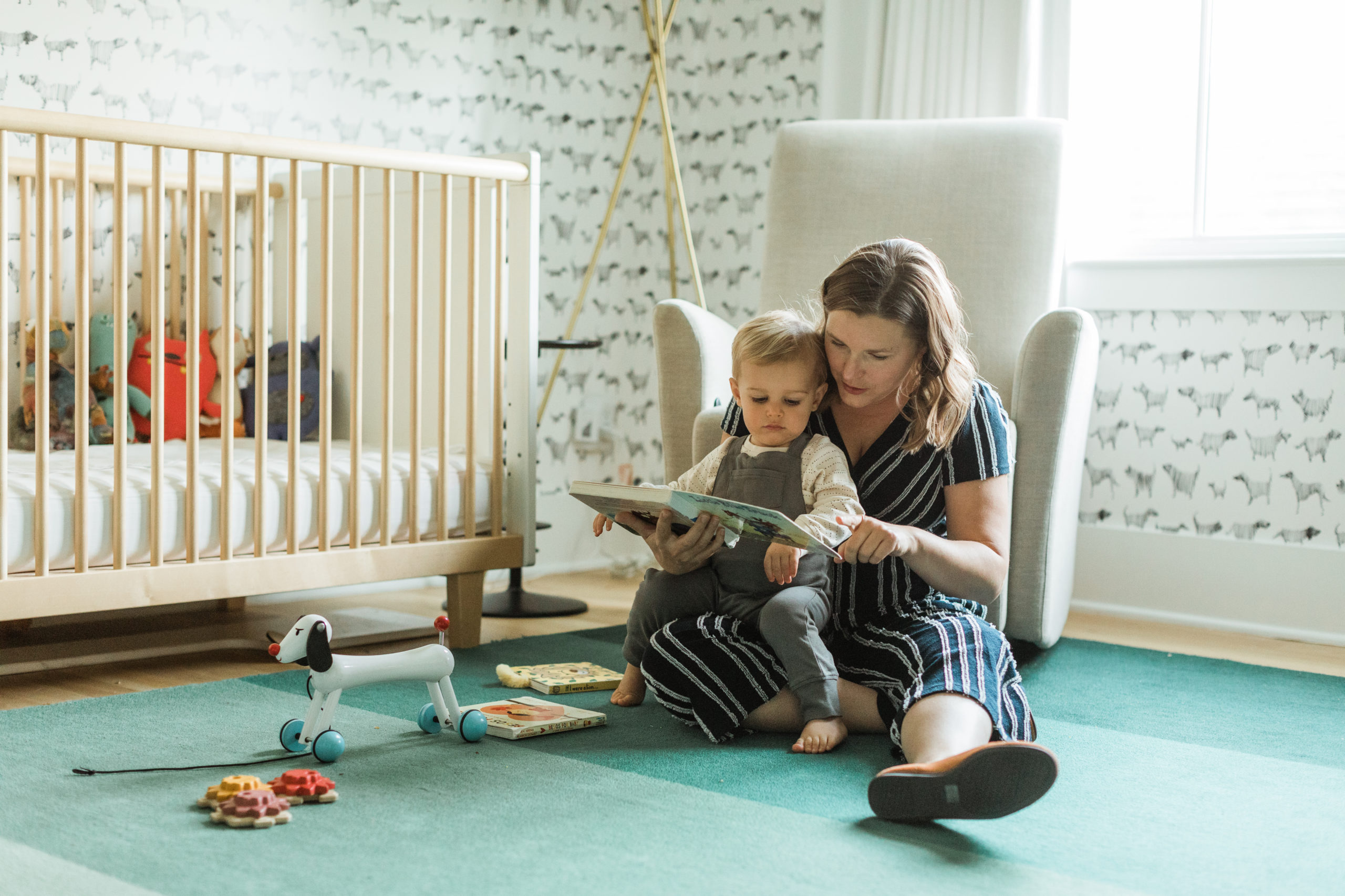Mama and baby boy sitting on the floor in little kid's bedroom reading a book. Animal print wall paper and light wooden furniture. Minimal aesthetic. Mama in black with white stripes romper.