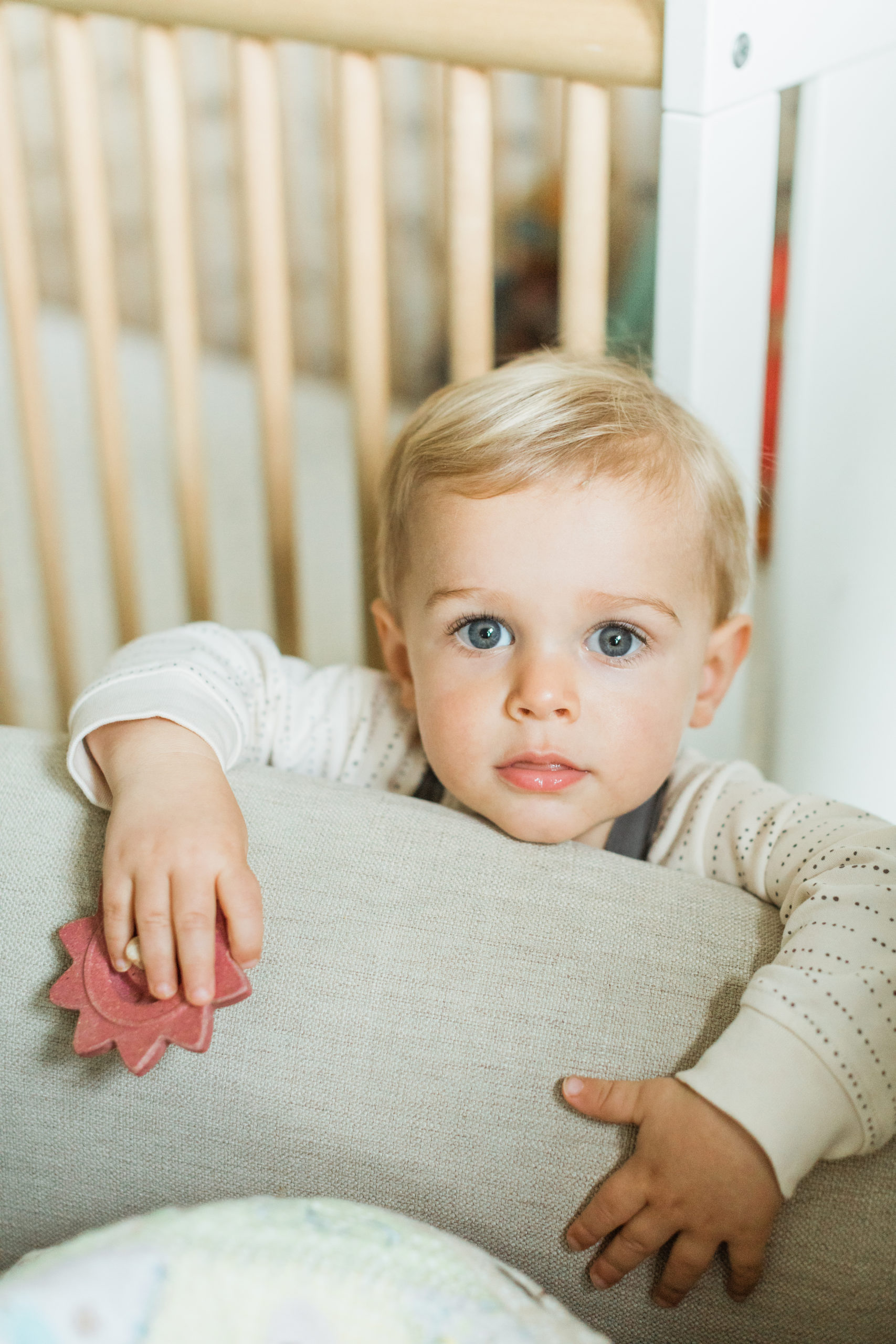 One year old baby boy with blue eyes staring at camera. Little boy leaning on cream couch wearing grey overalls and cream long sleeve.