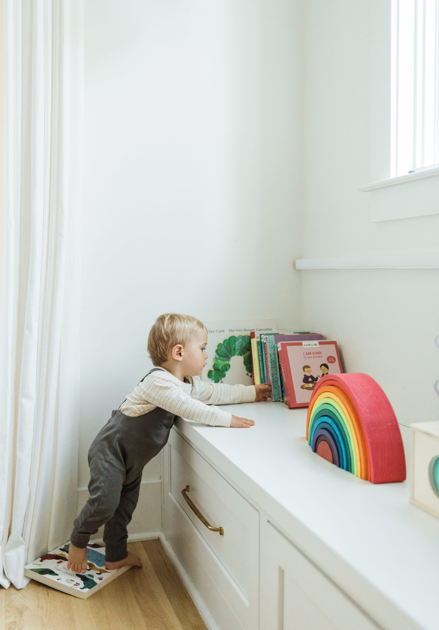One year old baby boy reaching for books in his bedroom. Rainbow toy, the very hungry caterpillar... Little boy wearing grey overalls and cream long sleeve with little dot details.