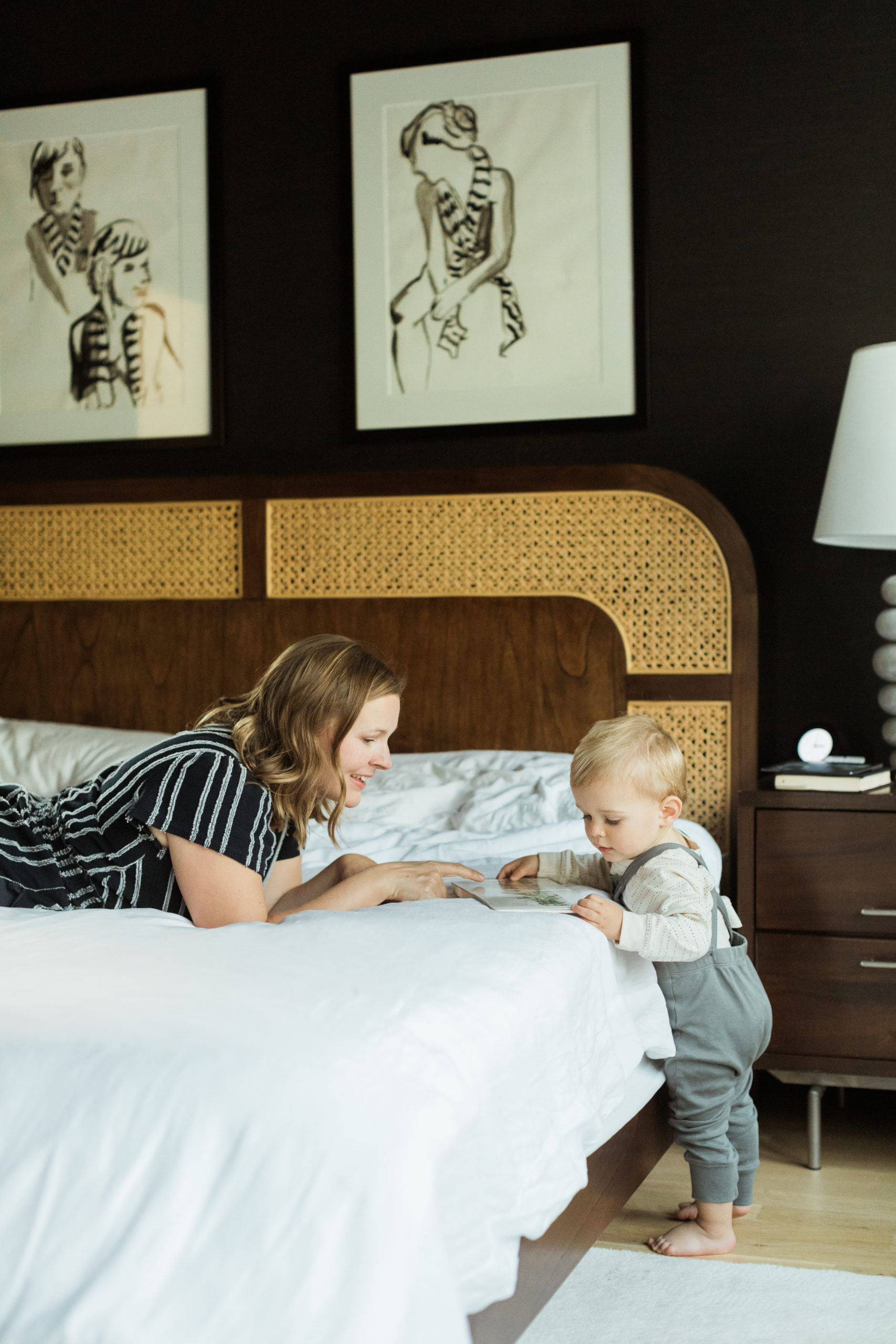 Photo of mama and her one year old baby boy. Mama is laying on her stomach in bed and little boy standing on edge of bed. Both mama and baby boy looking at book. Bedroom wall is black and headboard is wood. Beautiful minimal modern home decor.