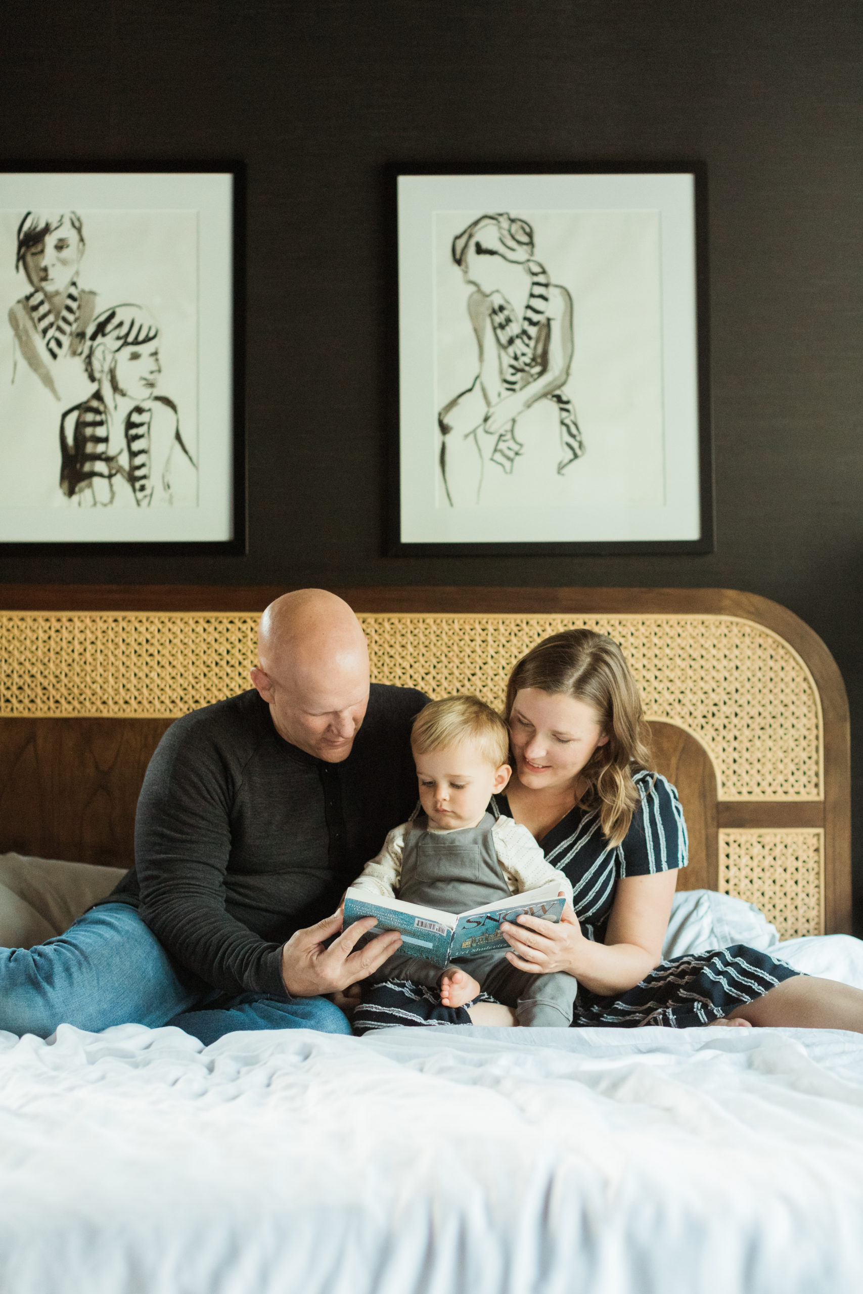 Mama, dad and baby boy sitting on bed reading a book. Black bedroom wall. Minimal aesthetic. Mama in black with white stripes romper.