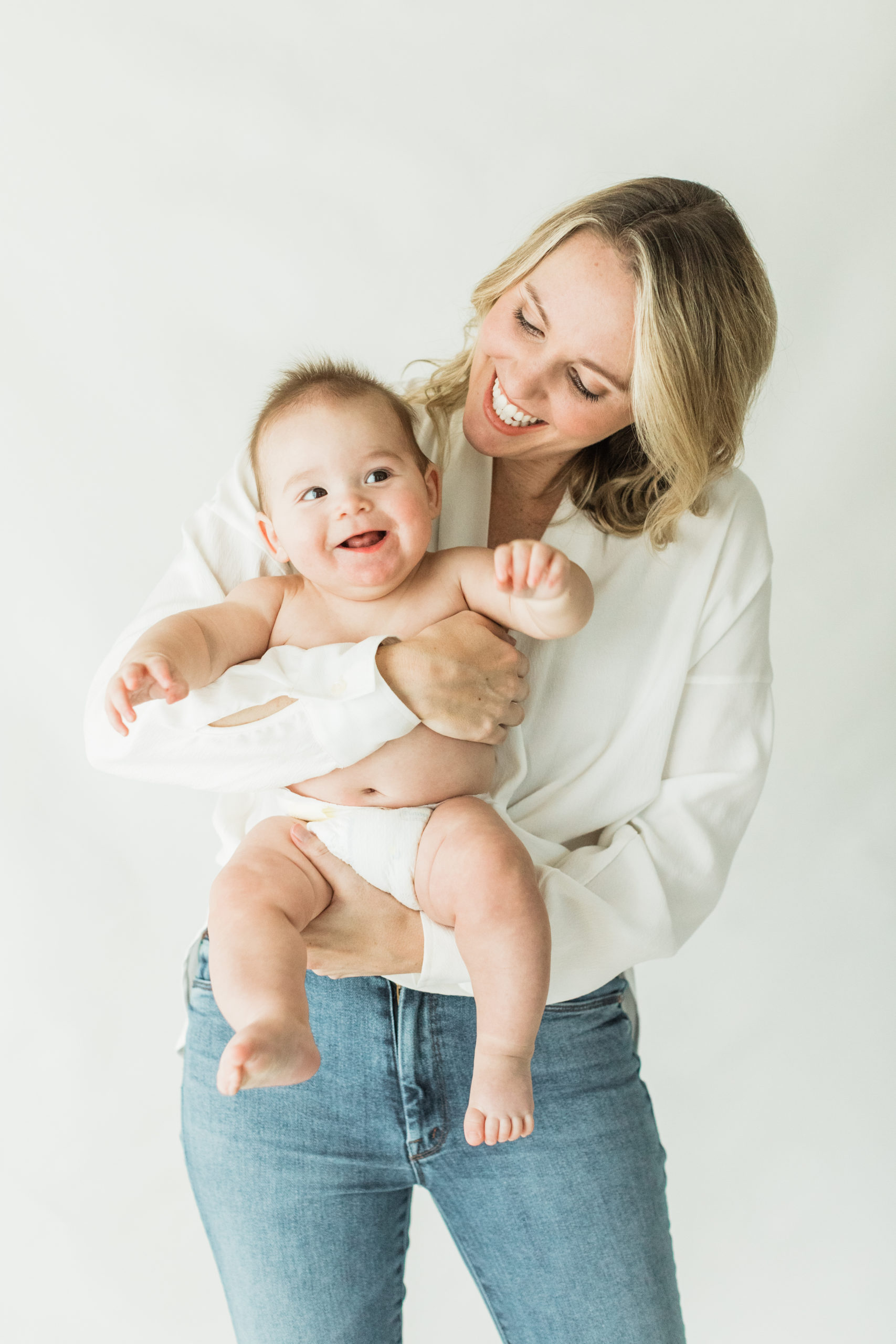 Mom holding her 6 month old baby boy. Baby boy in diaper, smiling. Mama in white long sleeve shirt and blue jeans. Natural light photographer.