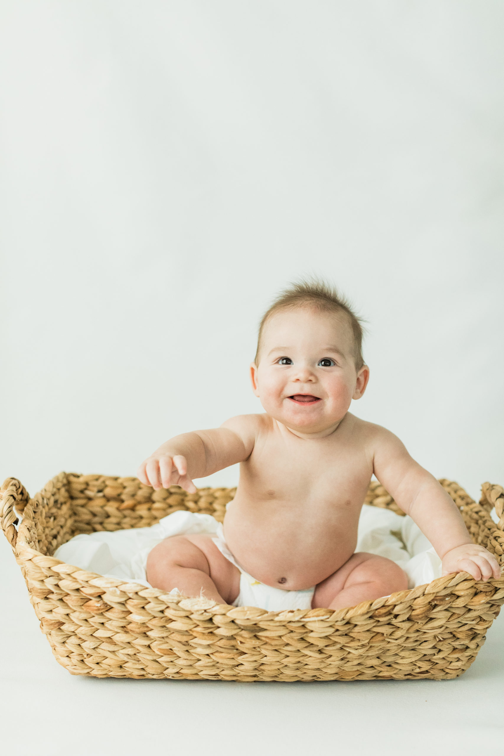 6 month old baby boy in diaper sitting in woven basket with white blanket smiling at camera. Natural light photographer.