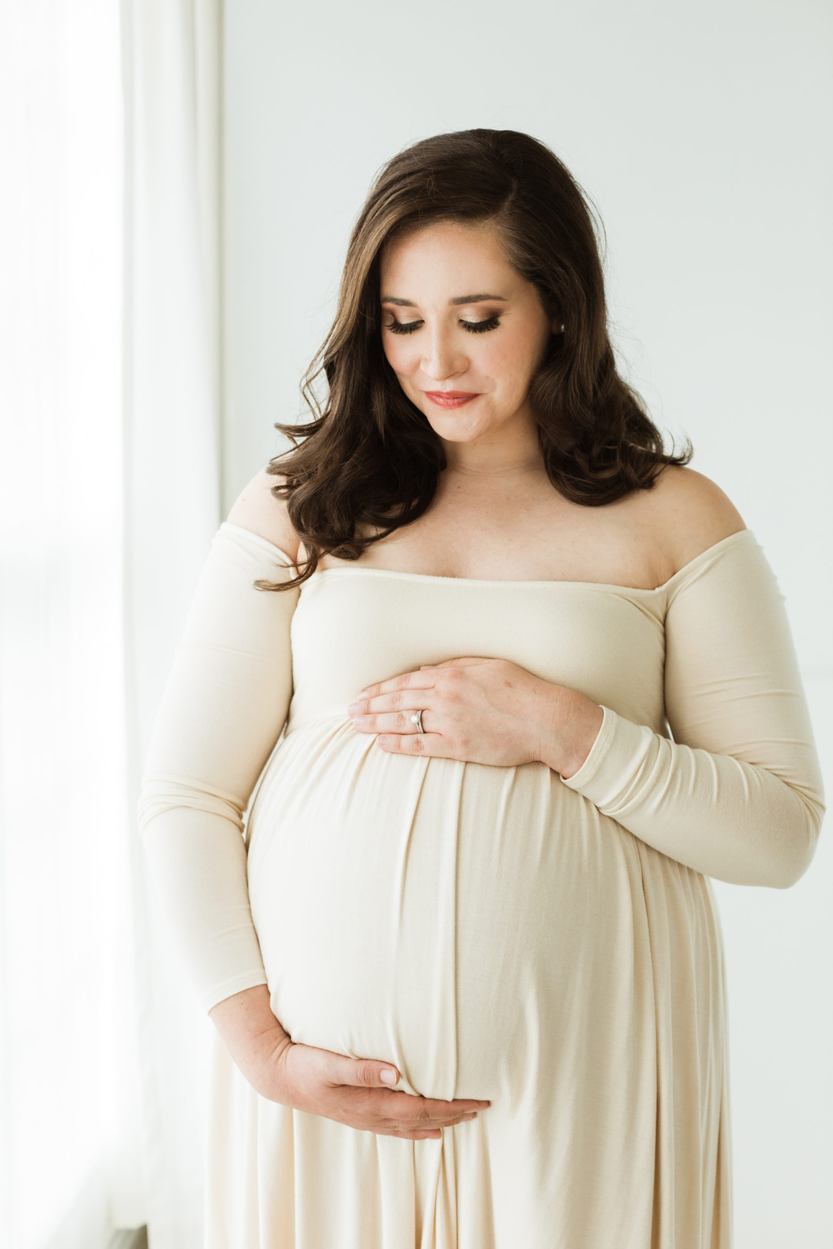 Neutral aesthetic. Photo of pregnant mama standing by window and looking down at her belly. Mama wearing a cream long sleeve off the shoulder maxi dress, both hands on her belly.
