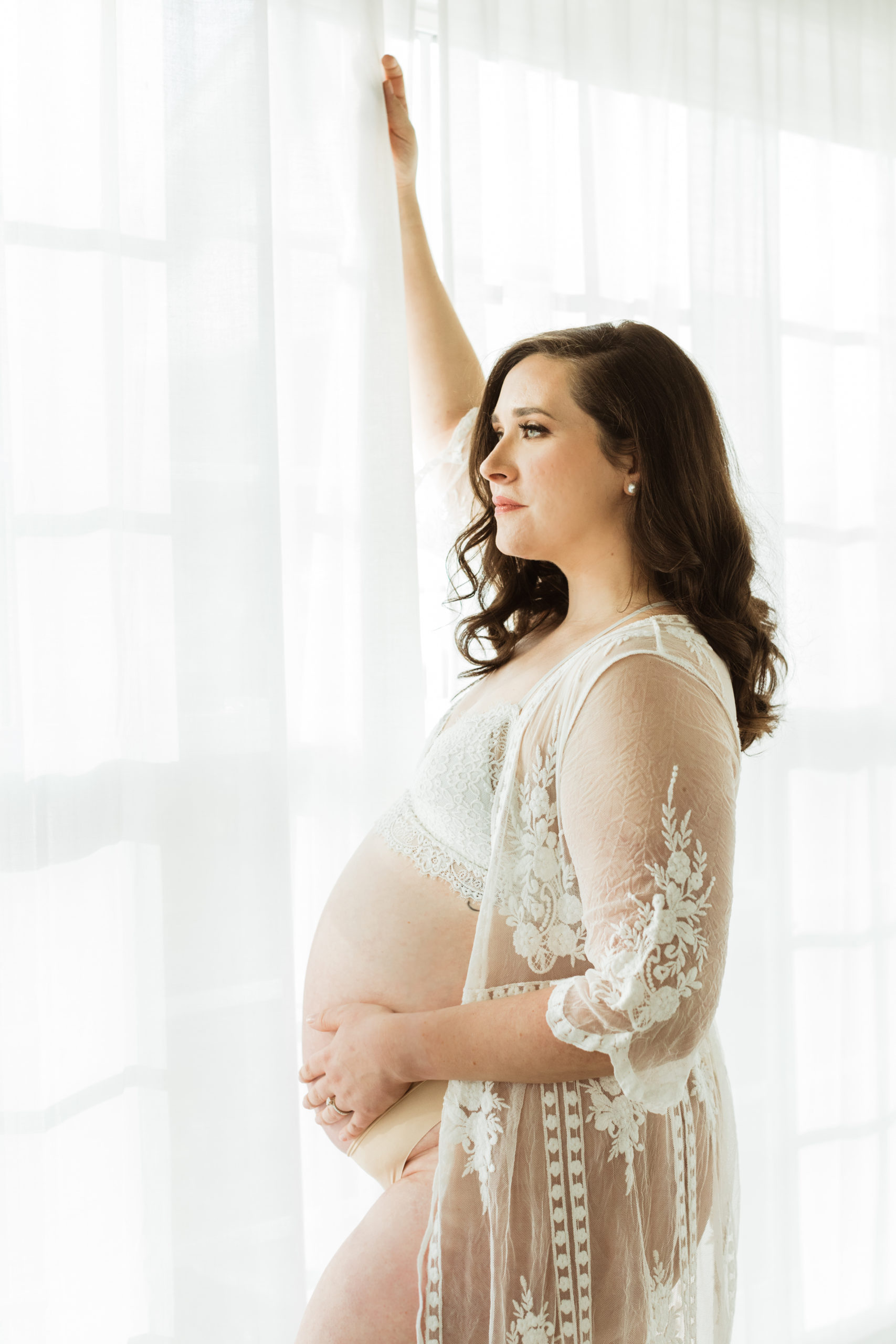 Pregnant mama looking outside a window wearing a see through nightgown with belly exposed. Indoor natural light photography.