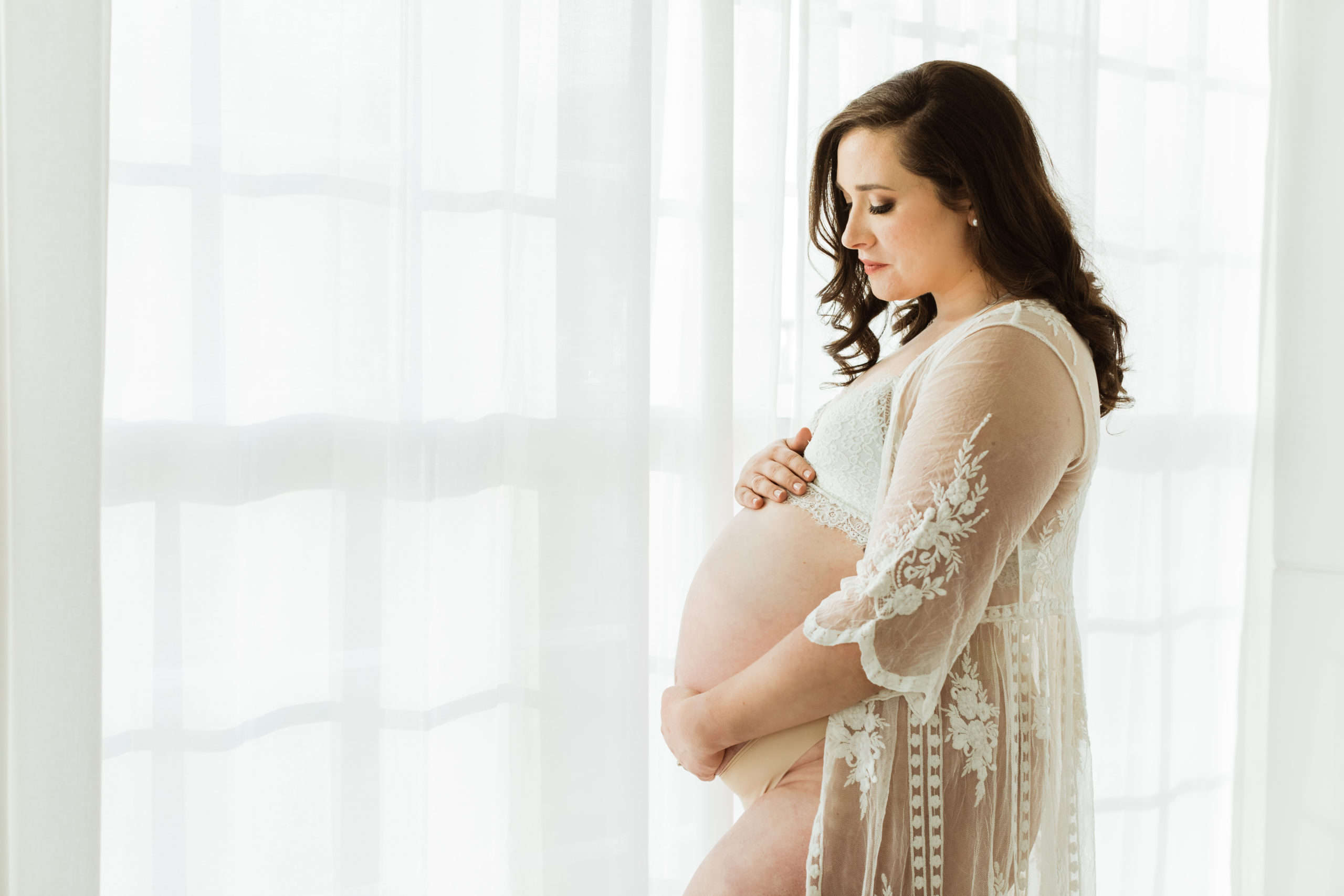 Pregnant mama looking at belly near window wearing a see through nightgown with belly exposed. Indoor natural light photography.