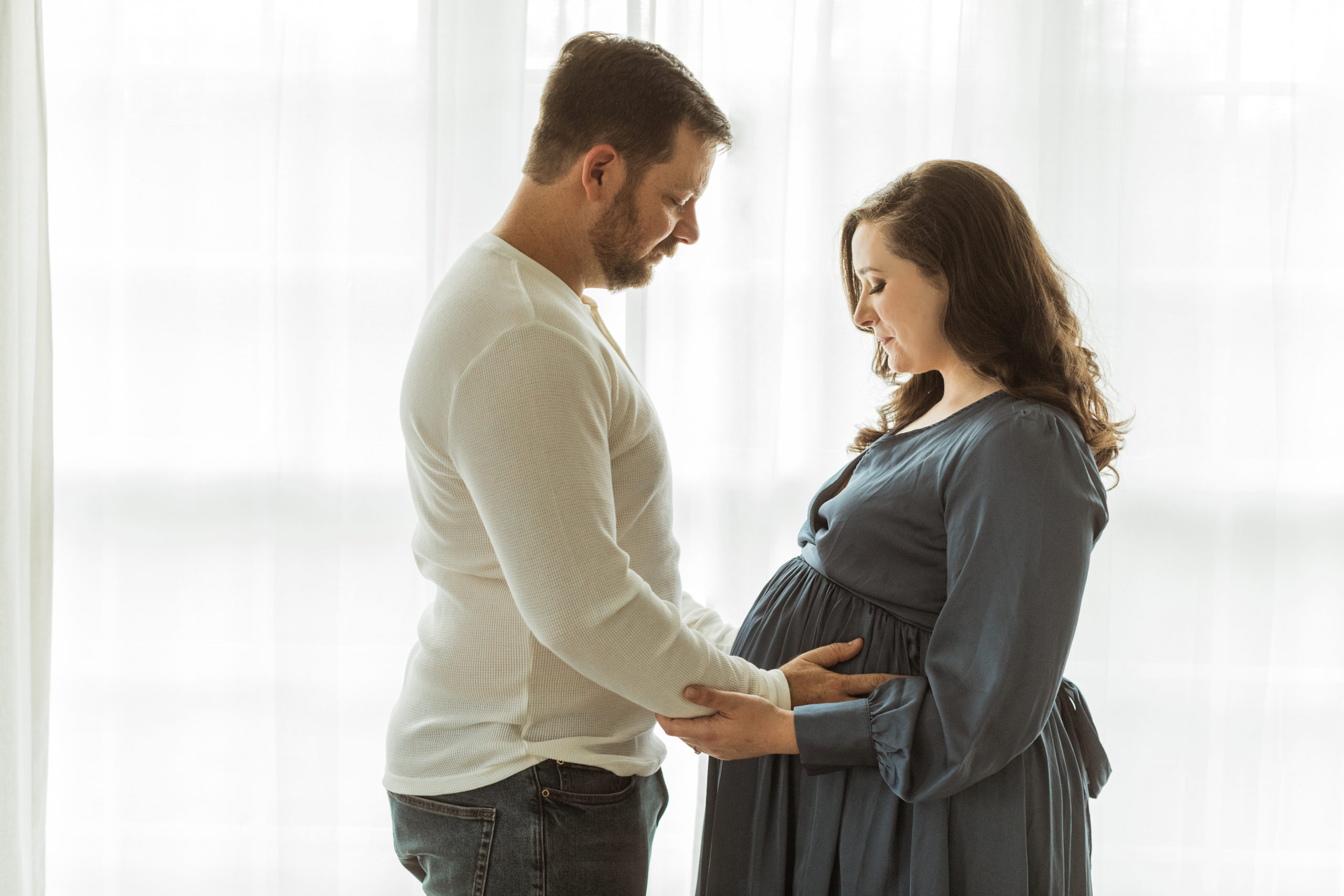 Mother and father. Husband holding wife's pregnant belly, wife's hands on her belly. Mama wearing silk blue long sleeve maxi dress. Man wearing long sleeve beige top and jeans. 