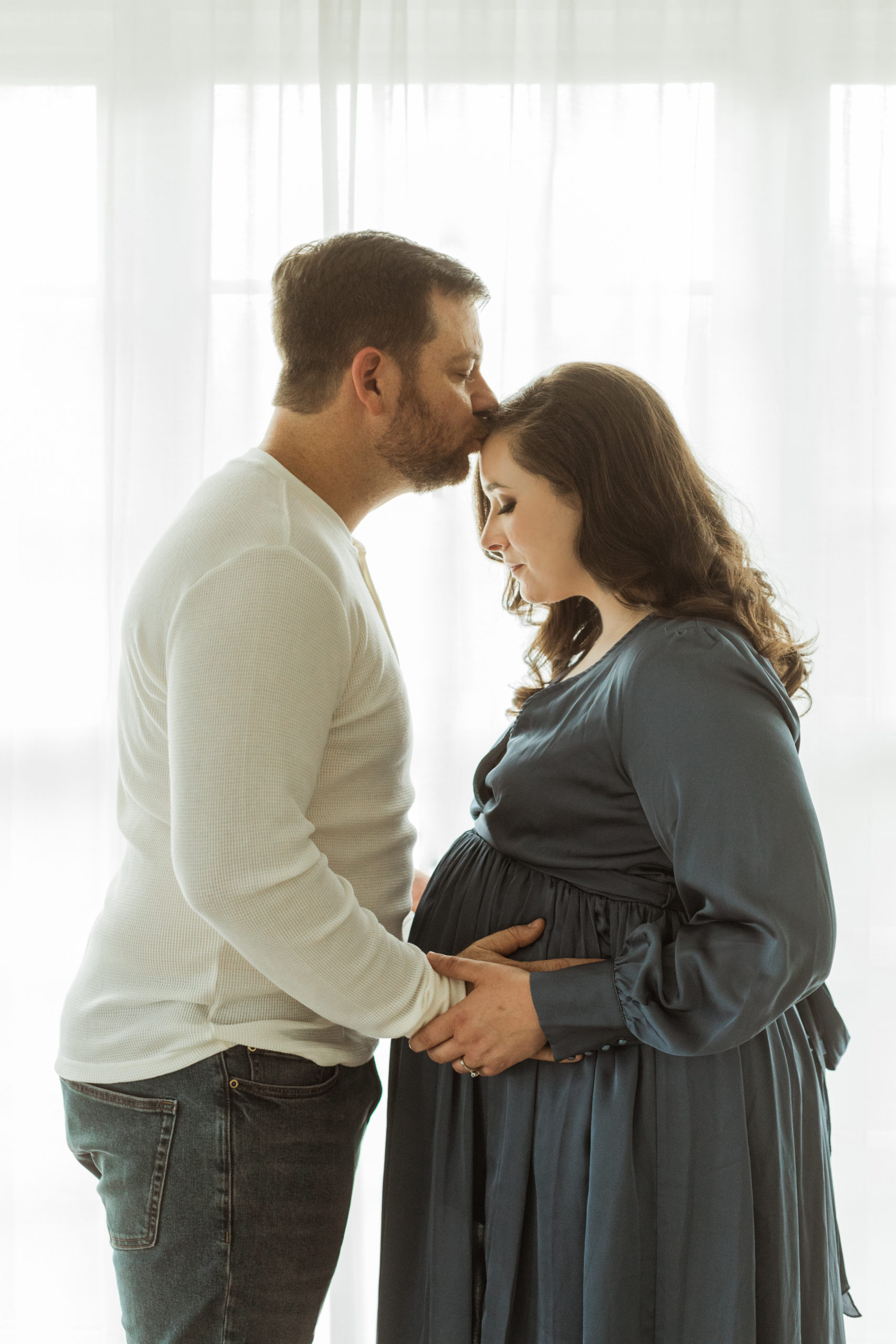 Mother and father. Husband holding wife's pregnant belly, wife's hands on her belly. Mama wearing silk blue long sleeve maxi dress. Man wearing long sleeve beige top and jeans. Husband kissing wife's forehead.