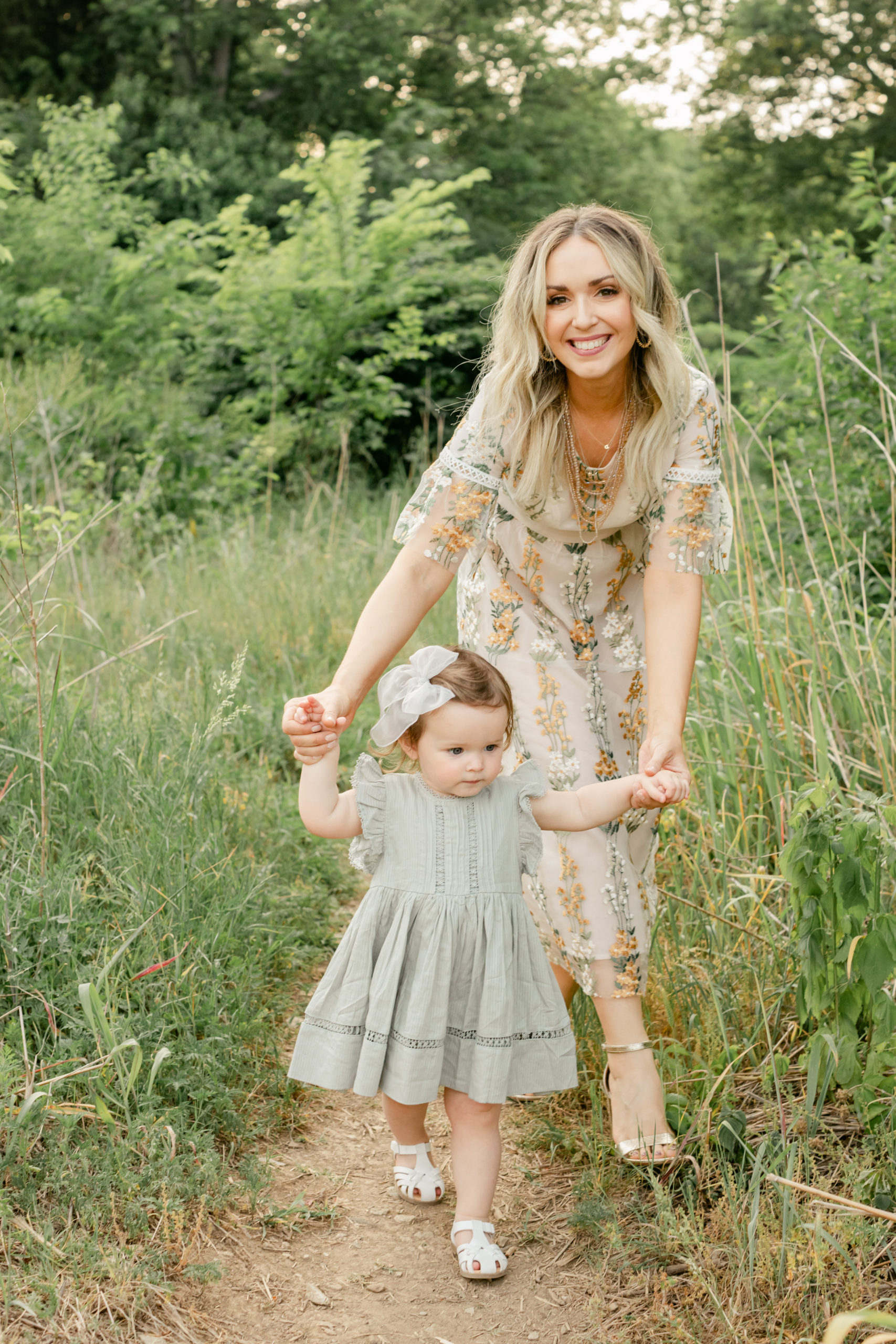 Photo of professional hair and makeup artist from Nashville outdoors with her daughter. Outdoor photo of mama and baby girl. Mama is holding daughters arms while they walk. Smiling.