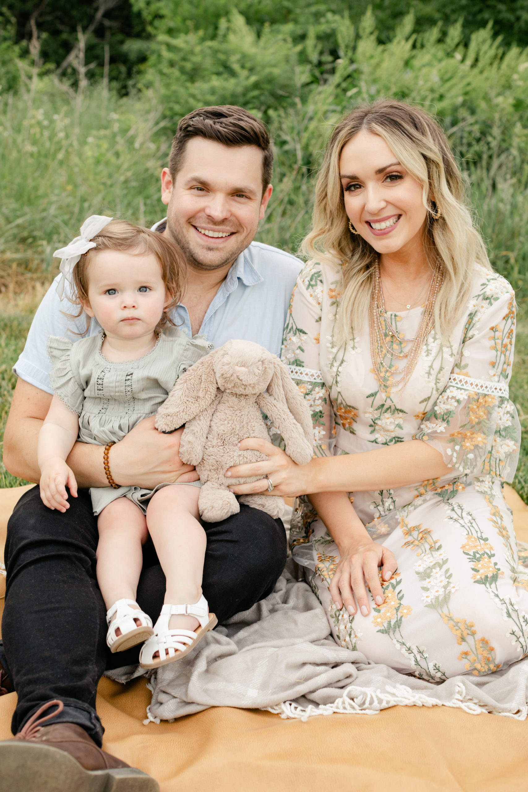Outdoor photo of family of three. Mom, dad and their baby girl sitting on blanket on the grass. Family smiling towards camera. Mama wearing lace details floral embroidered maxi dress. Dad wearing black pants and light blue collar button up. Baby girl wearing white bow in hair and sage green dress holding her stuffed bunny.