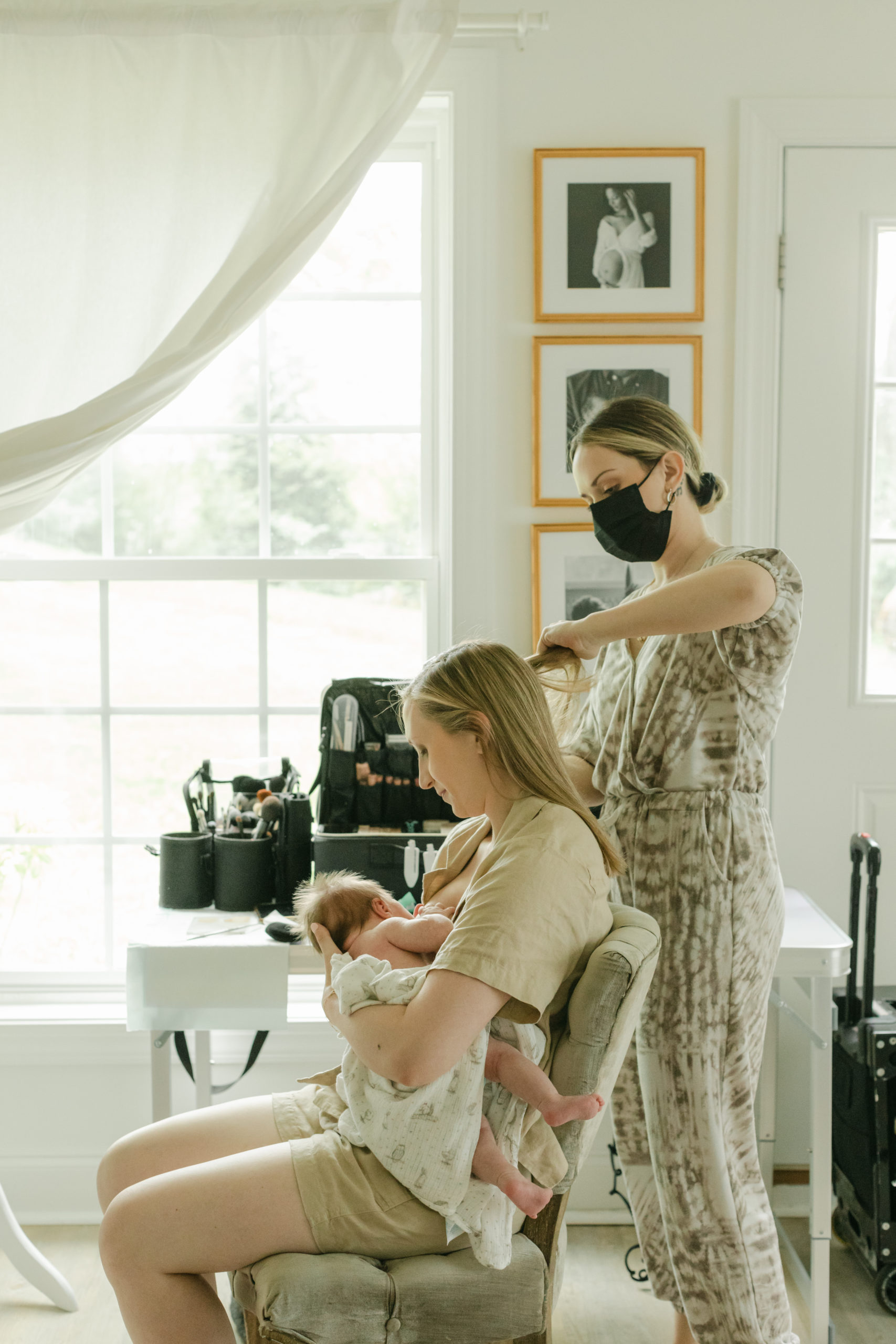 Professional hair and makeup artist doing makeup on client in preparation for a family photo shoot. Hair and makeup artist wearing a face mask, working on clients hair while mama is breastfeeding her baby.
