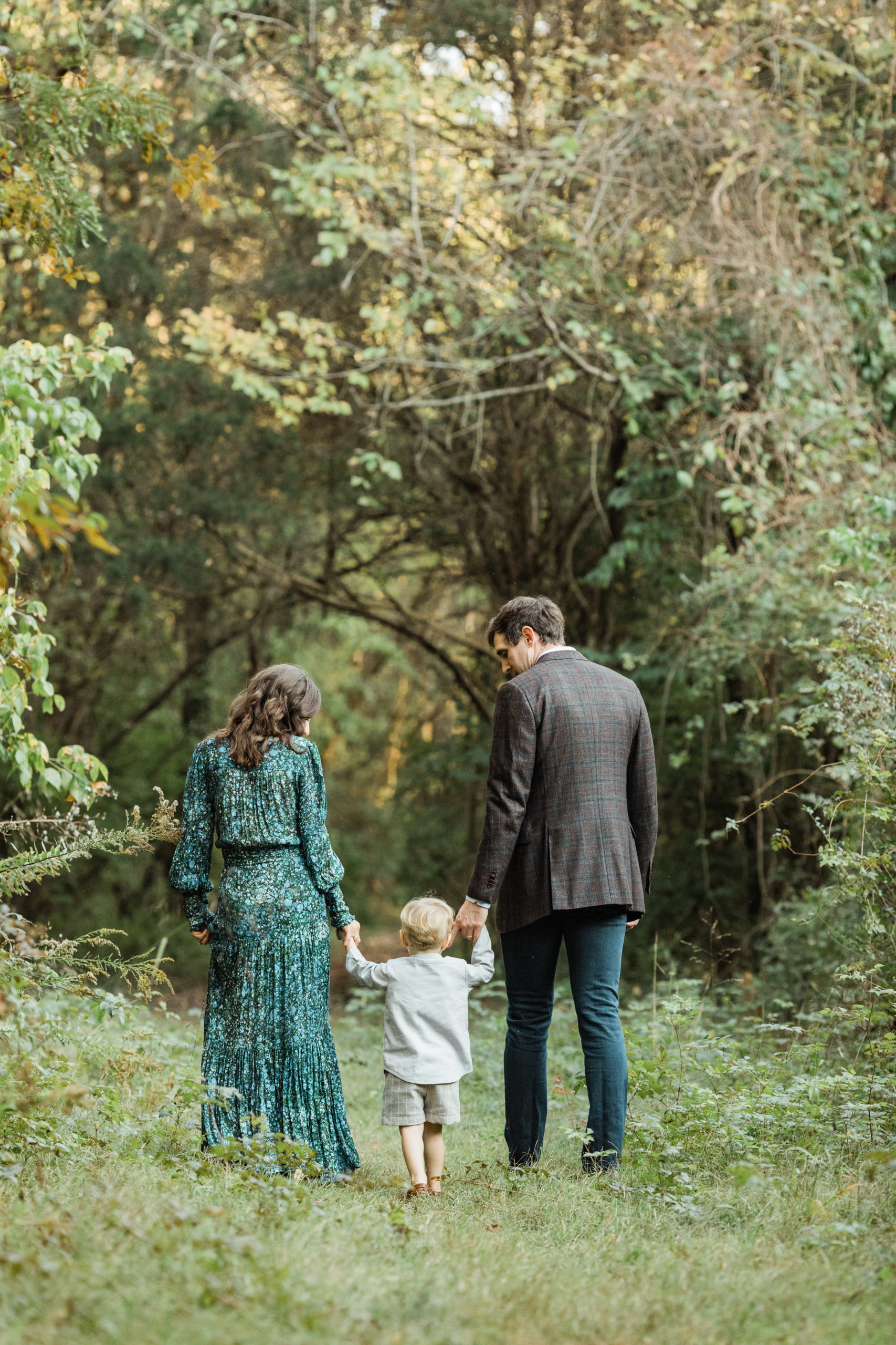 Photo of family of three outdoors for fall family session. Dad wearing plaid blazer, mom wearing green and blue long sleeve maxi dress, baby boy wearing light blue button up. Family of three holding hands walking away from camera.
