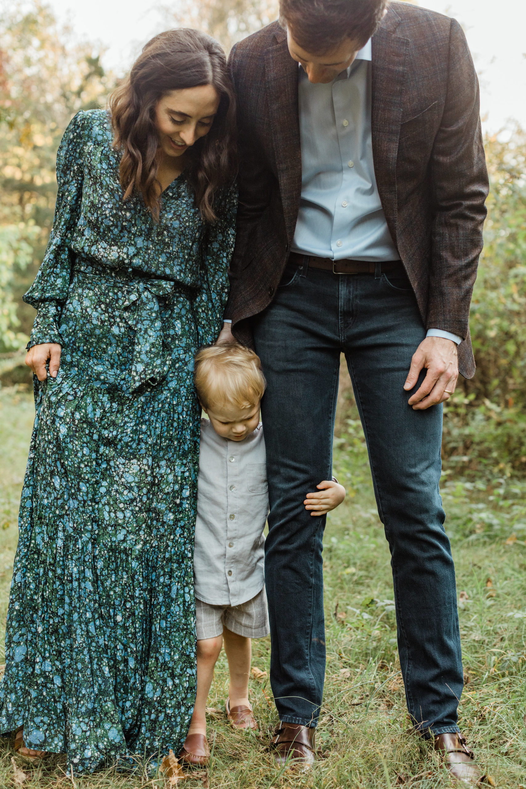 Outdoor photo of family of three. Mama wearing blue and green long sleeve maxi dress, dad wearing brown blazer, jeans and light blue button up. Little boy standing between both parents' legs.