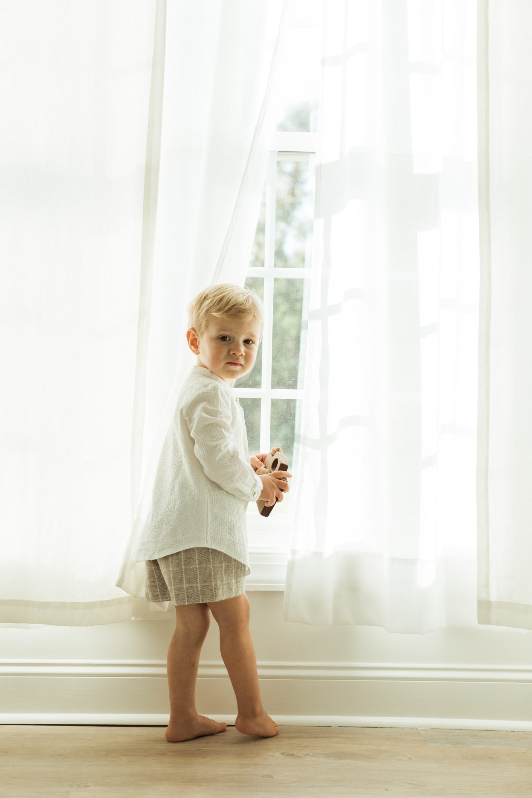 Little boy standing near window. White curtains and white walls. Little boy wearing tan shorts and white button up.