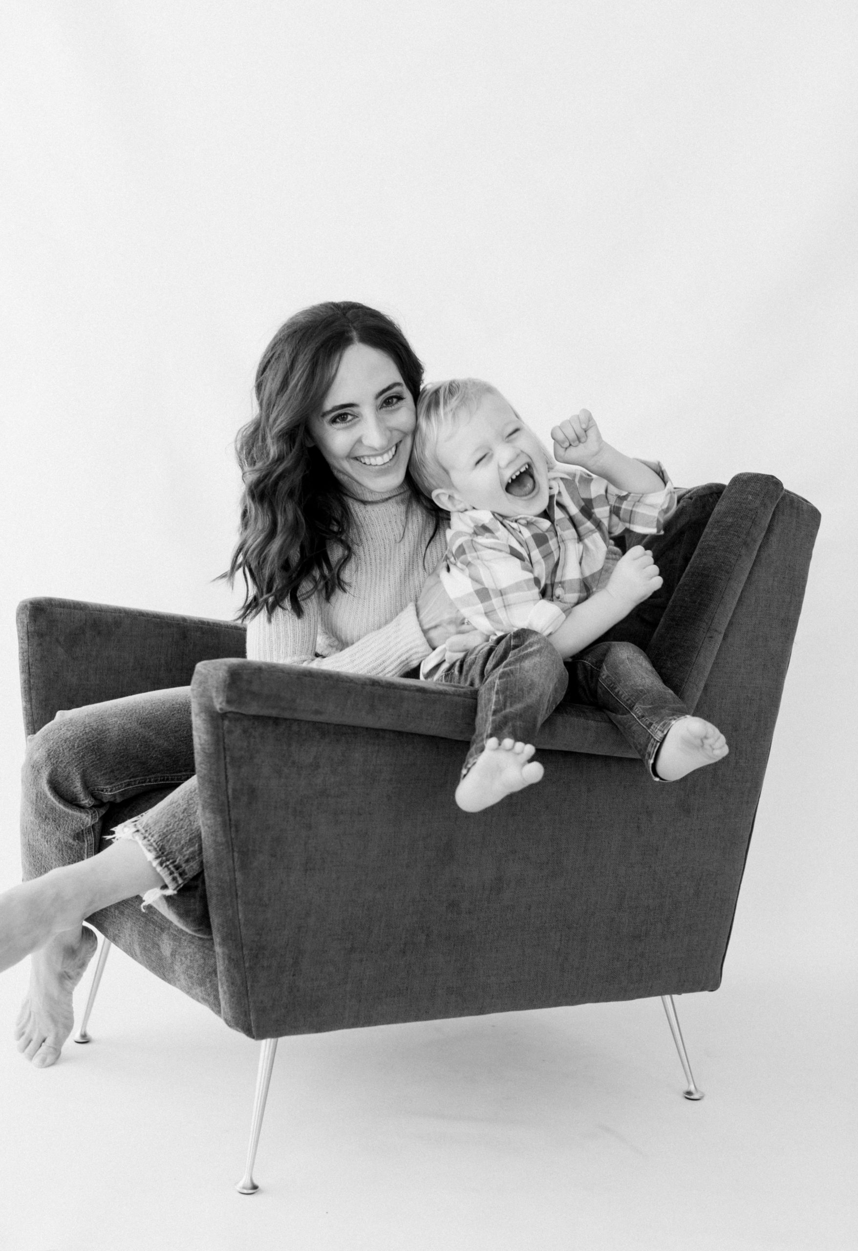 black and white photo of mama and baby boy sitting on velvet sofa chair. Little boy wearing jeans and plaid shirt. Mama tickling son.