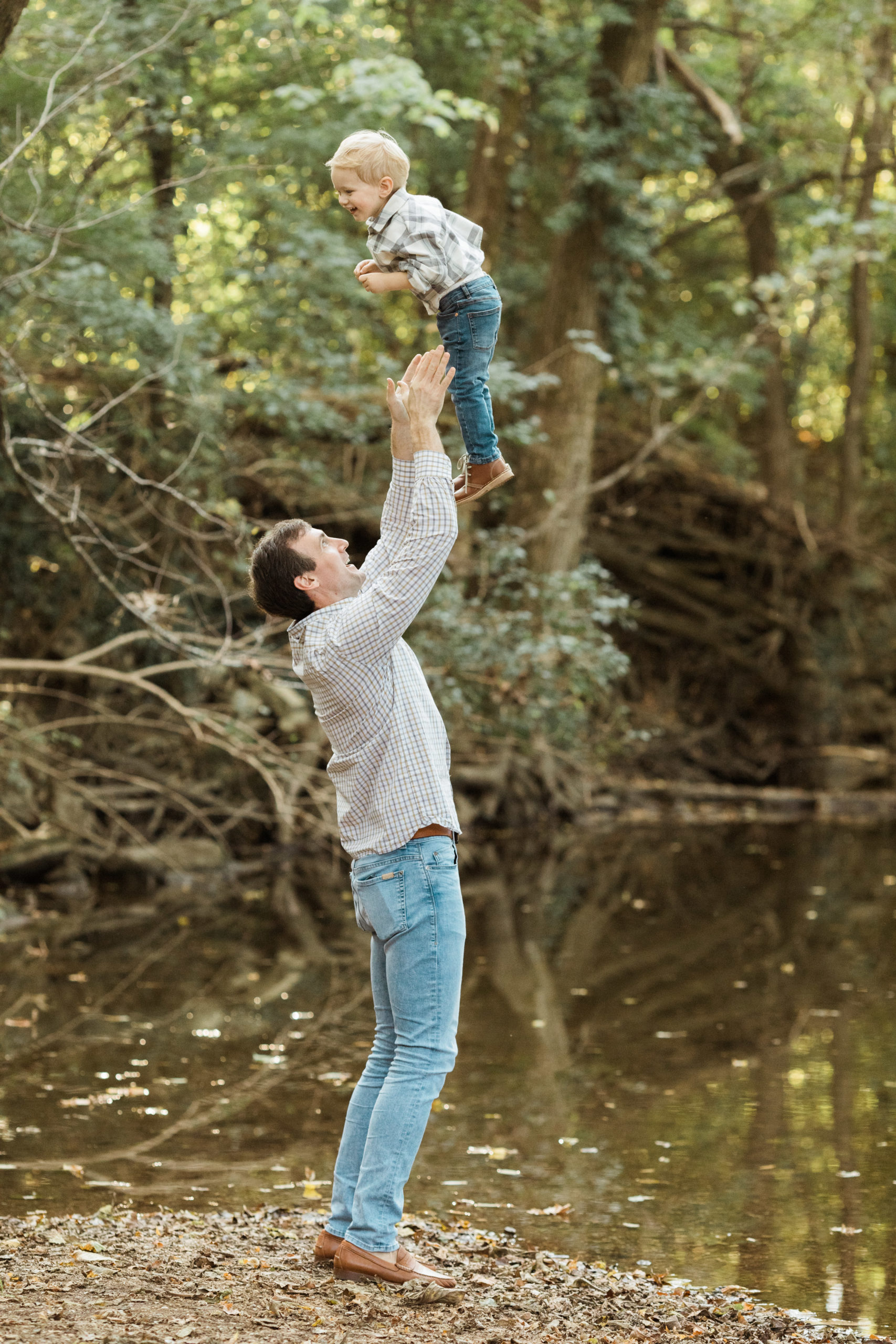 Outdoor photo of dad holding his son up in the air. Near a pond of water.