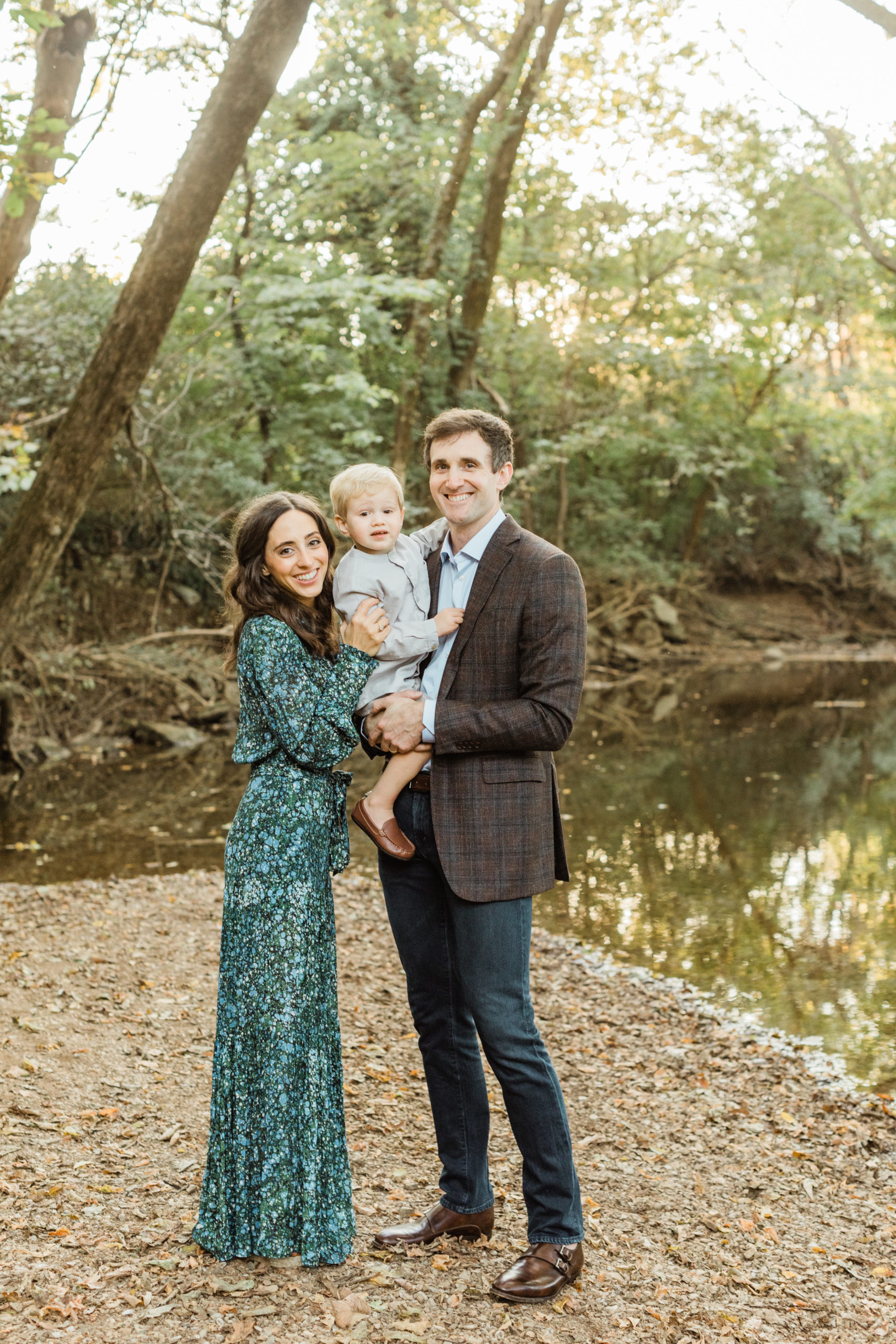 Photo of family of three outdoors for fall family session. Dad wearing plaid blazer, mom wearing green and blue long sleeve maxi dress, baby boy wearing light blue button up. Family smiling at each other.