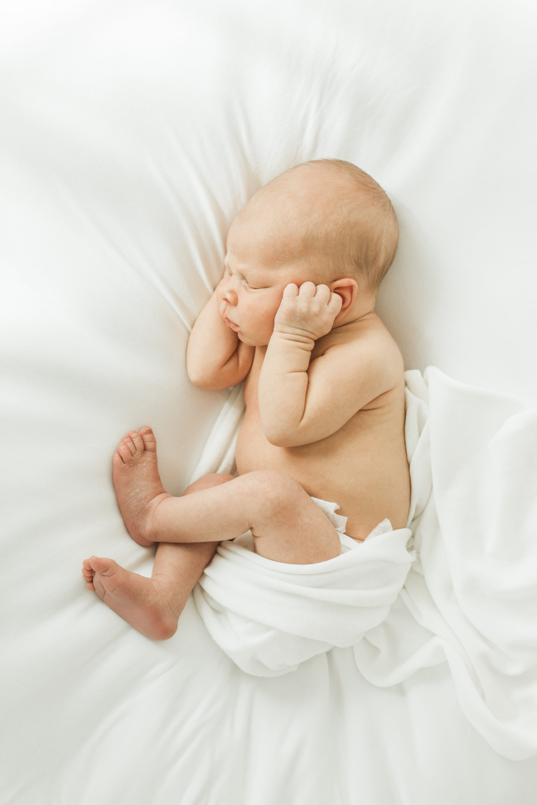 Newborn baby boy on white bed wrapped in white blanket. Baby's hands covering his ears.