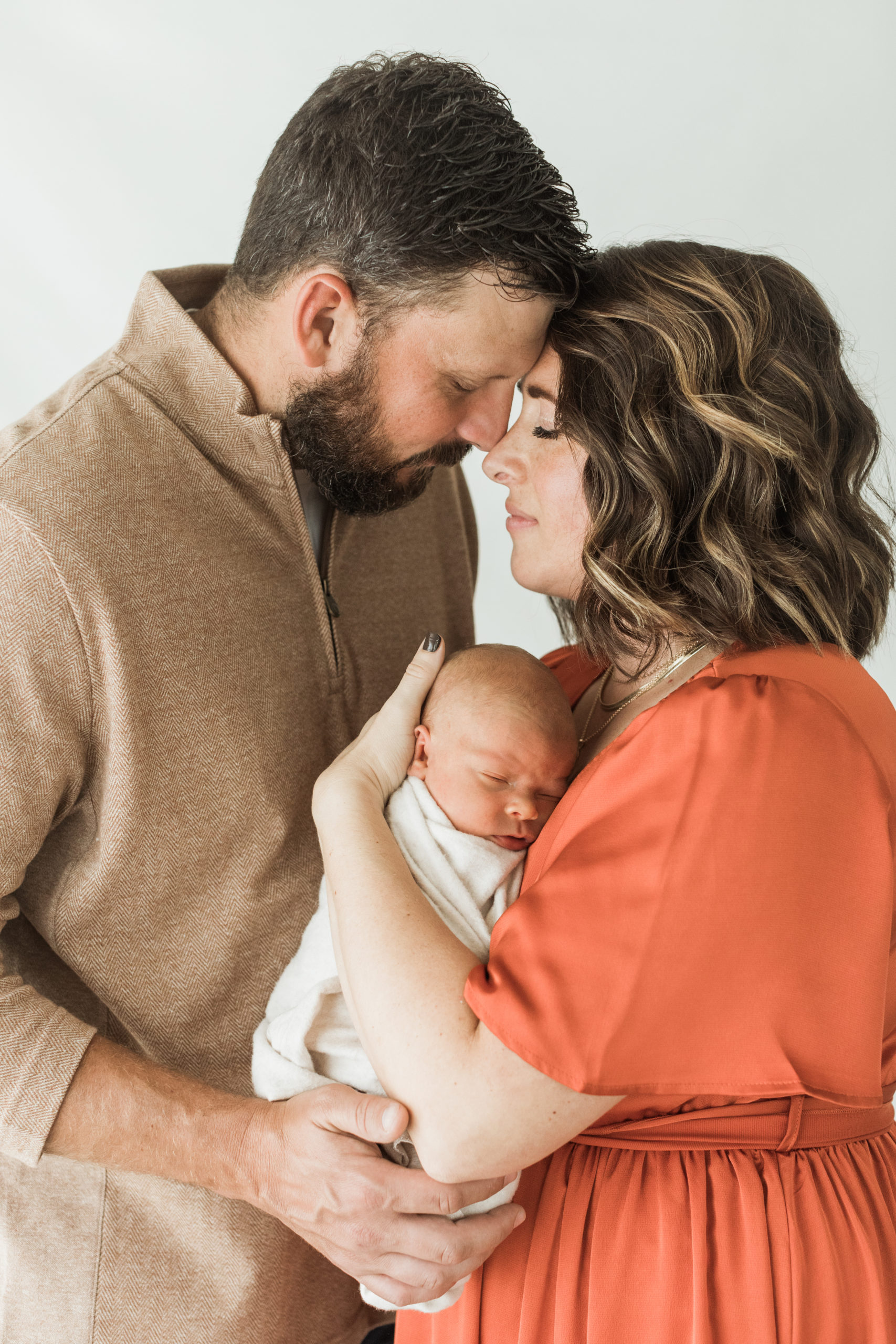 Photo of mom and dad with their newborn baby boy in mamas arms. Both parents holding baby wrapped in beige blanket and parents with their eyes closed and foreheads are touching.