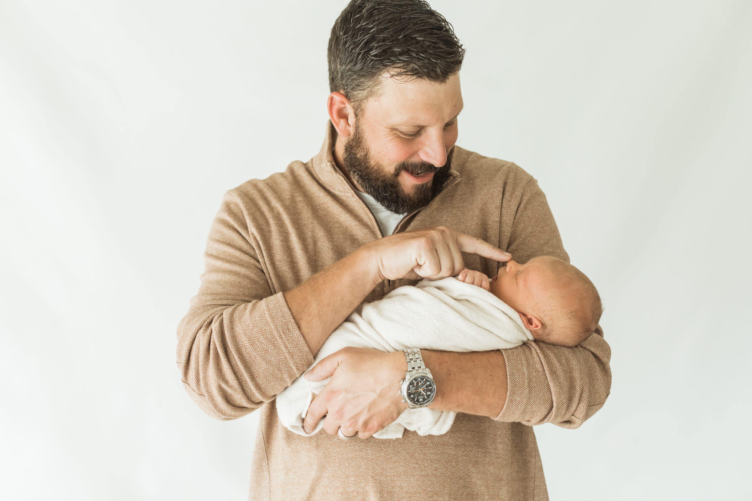 Dad holding his newborn baby boy in his arms. Baby wrapped in beige blanket. Dad touching baby's nose. Dad wearing tan sweater.