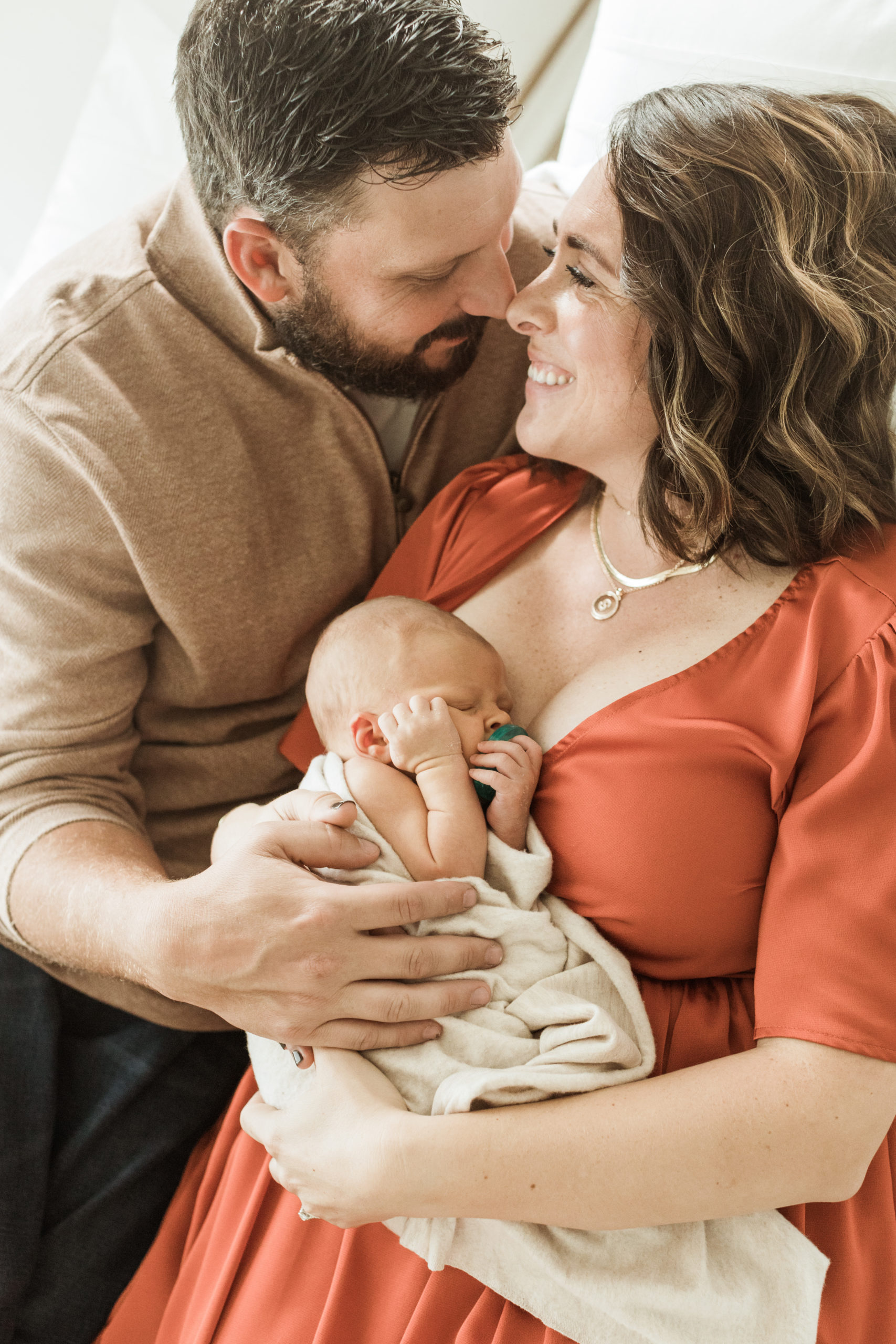 Photo of mom and dad with their newborn baby boy in mamas arms. Both parents are laying on bed, holding baby wrapped in beige blanket and parents looking at each other.