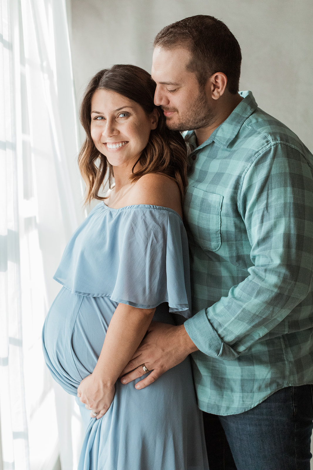 woman and man standing by window and smiling. pregnant mama wearing blue maternity dress.