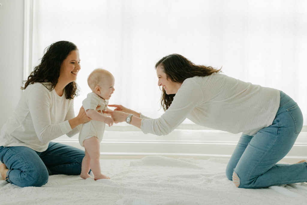 Photo of two mamas and their 6 month old baby boy. LGBTQ. Mamas wearing white long sleeves and blue jeans, baby boy standing up wearing white onesie.