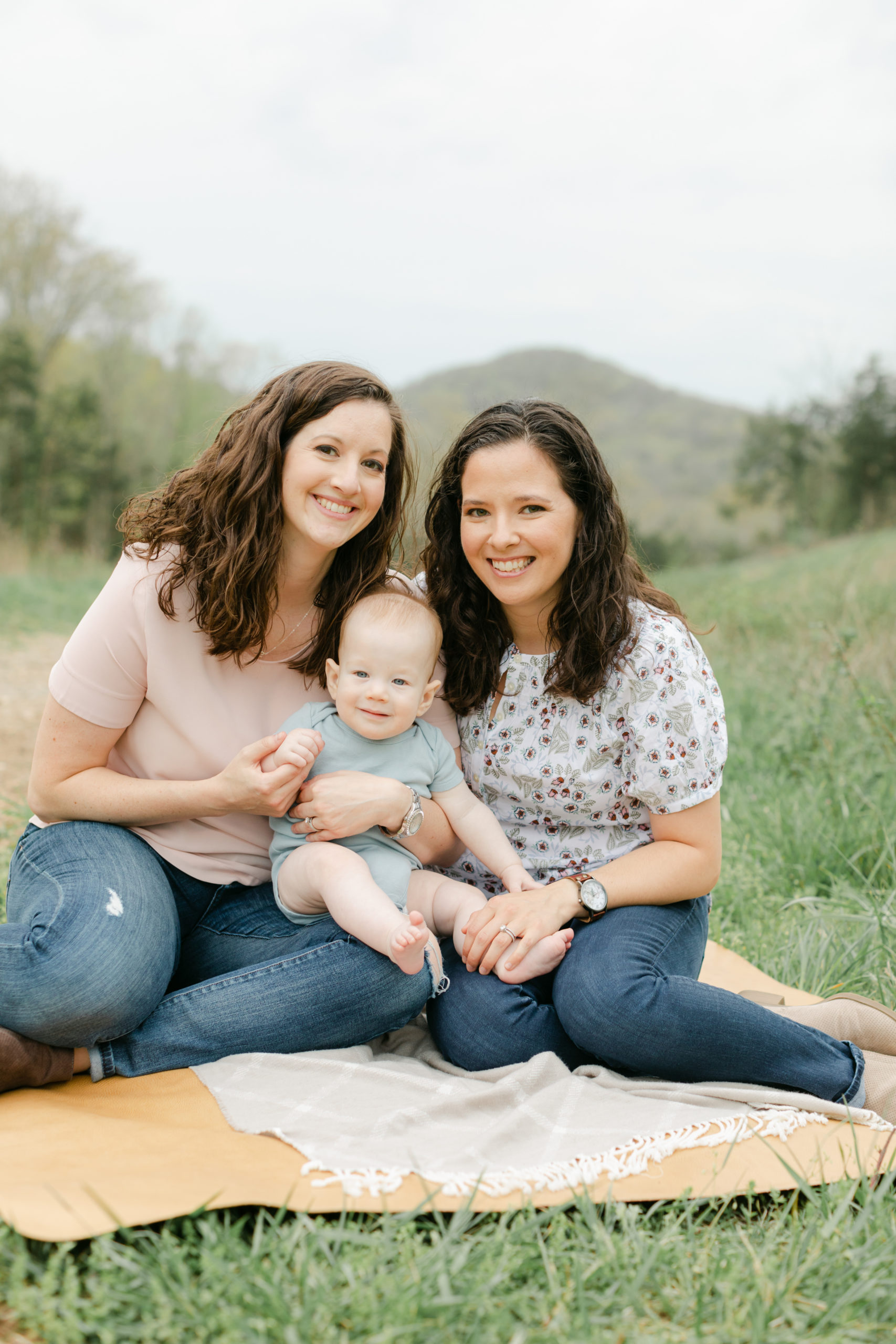 Outdoor photo of two mamas sitting on the grass, holding their 6 month old baby boy and smiling at camera.