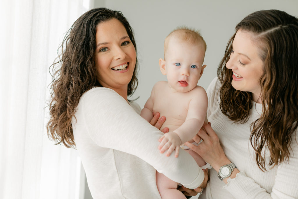 Photo of two mamas and their 6 month old baby boy. LGBTQ. Mamas wearing white long sleeves and blue jeans, baby boy in diaper.