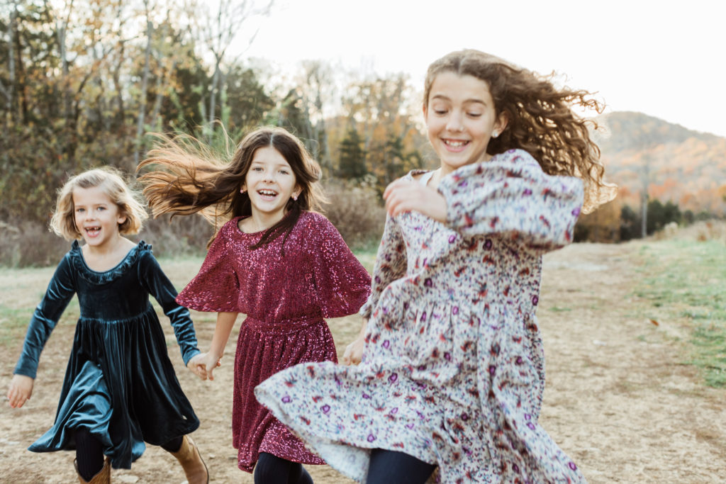 Three young sisters holding hands running, skipping and jumping around. Fall family photos.