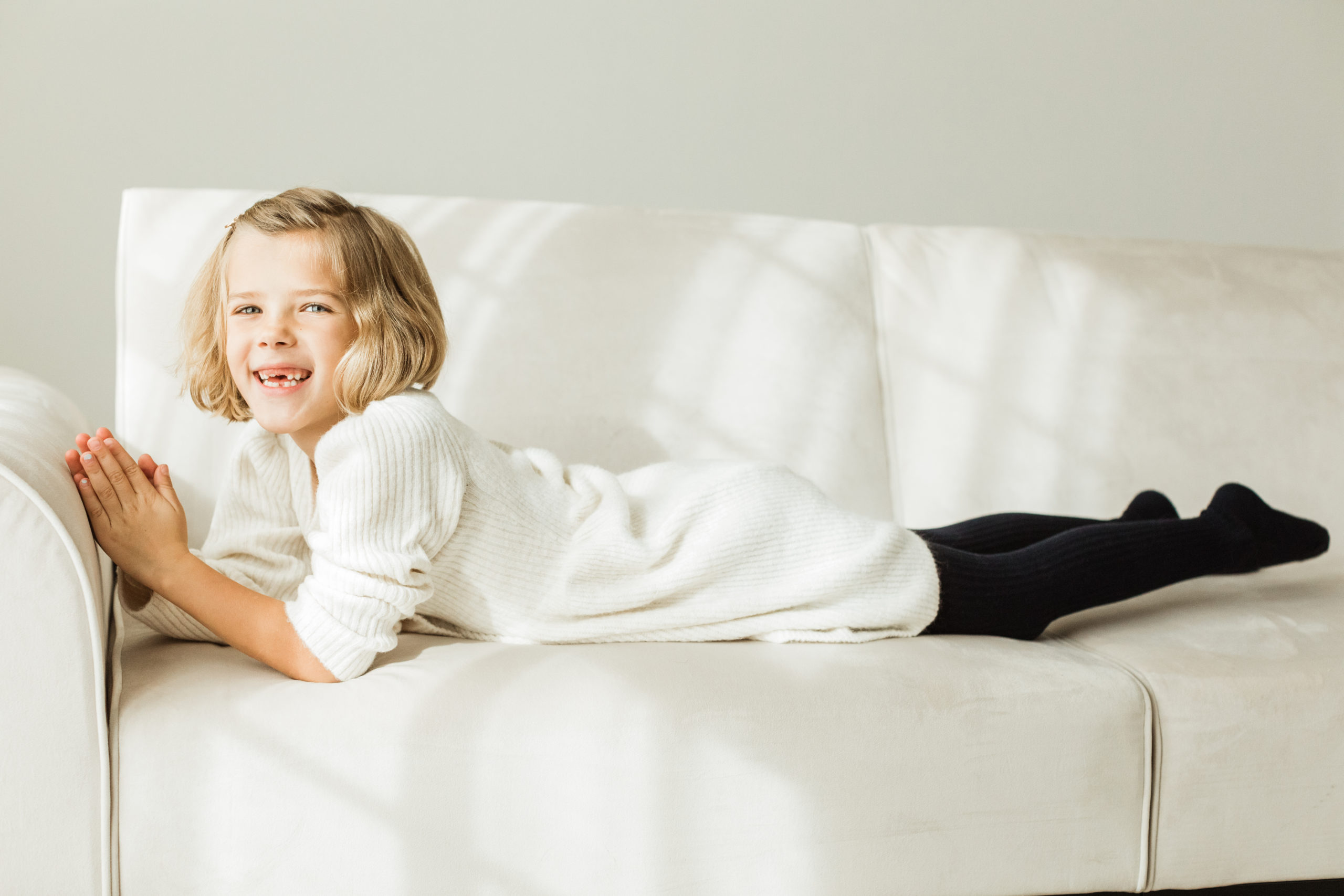 Young girl in white sweater dress and black tights smiling and laying on her belly on white couch.