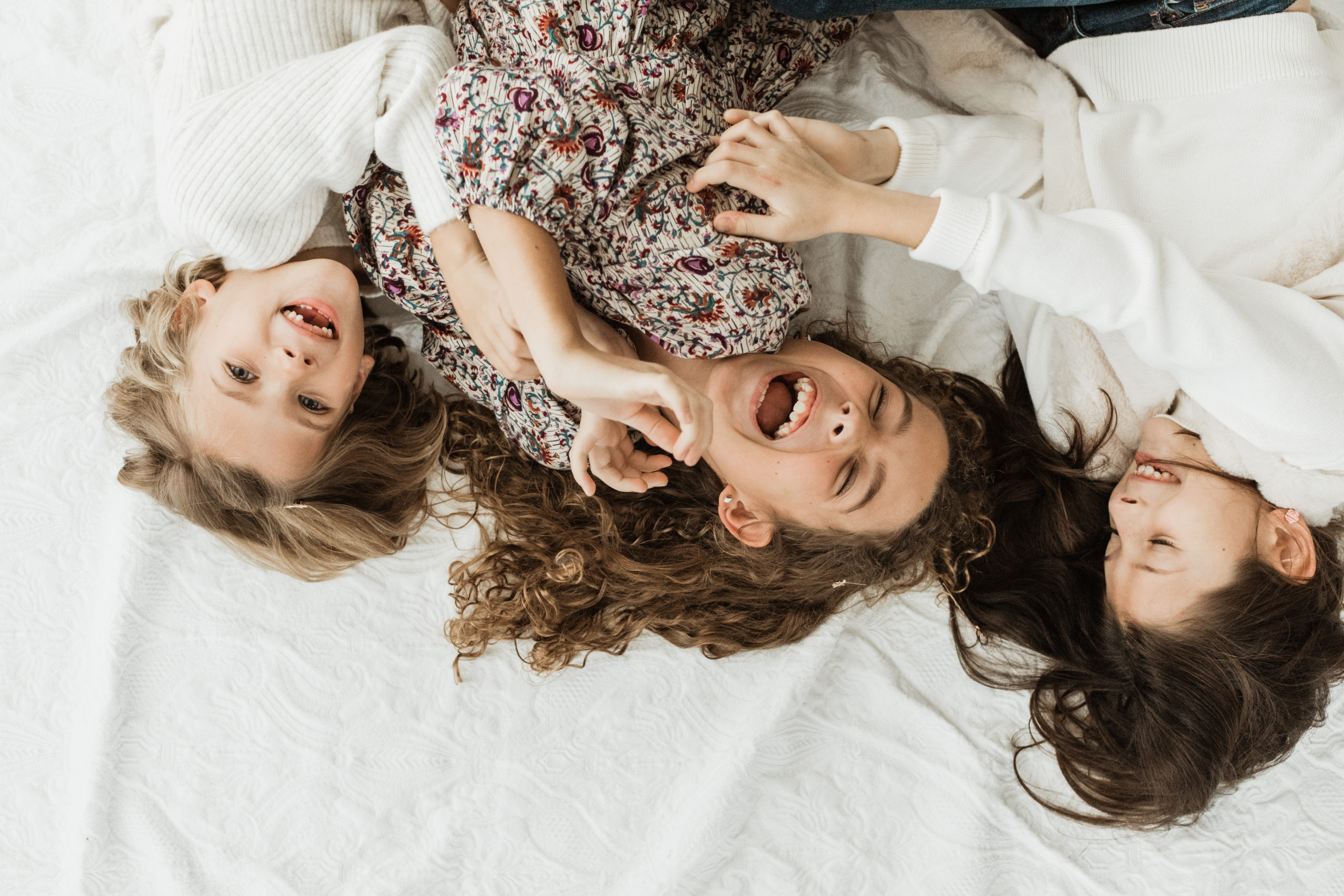 Three young sisters laughing and playing around, laying down on white sheet.