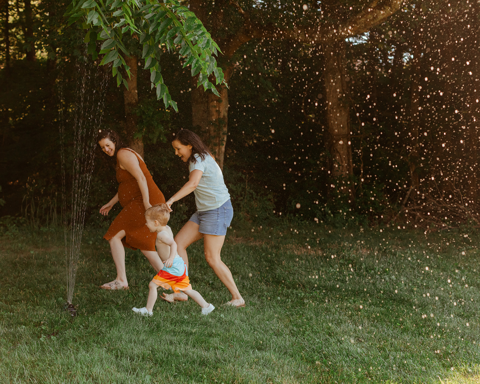 Moms running through sprinklers with son. Backyard summer family session in Nashville, TN