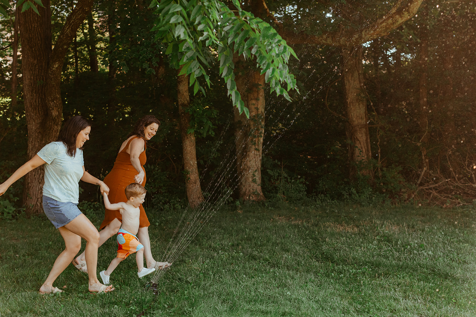 Moms running through sprinklers with son. Backyard summer family session in Nashville, TN