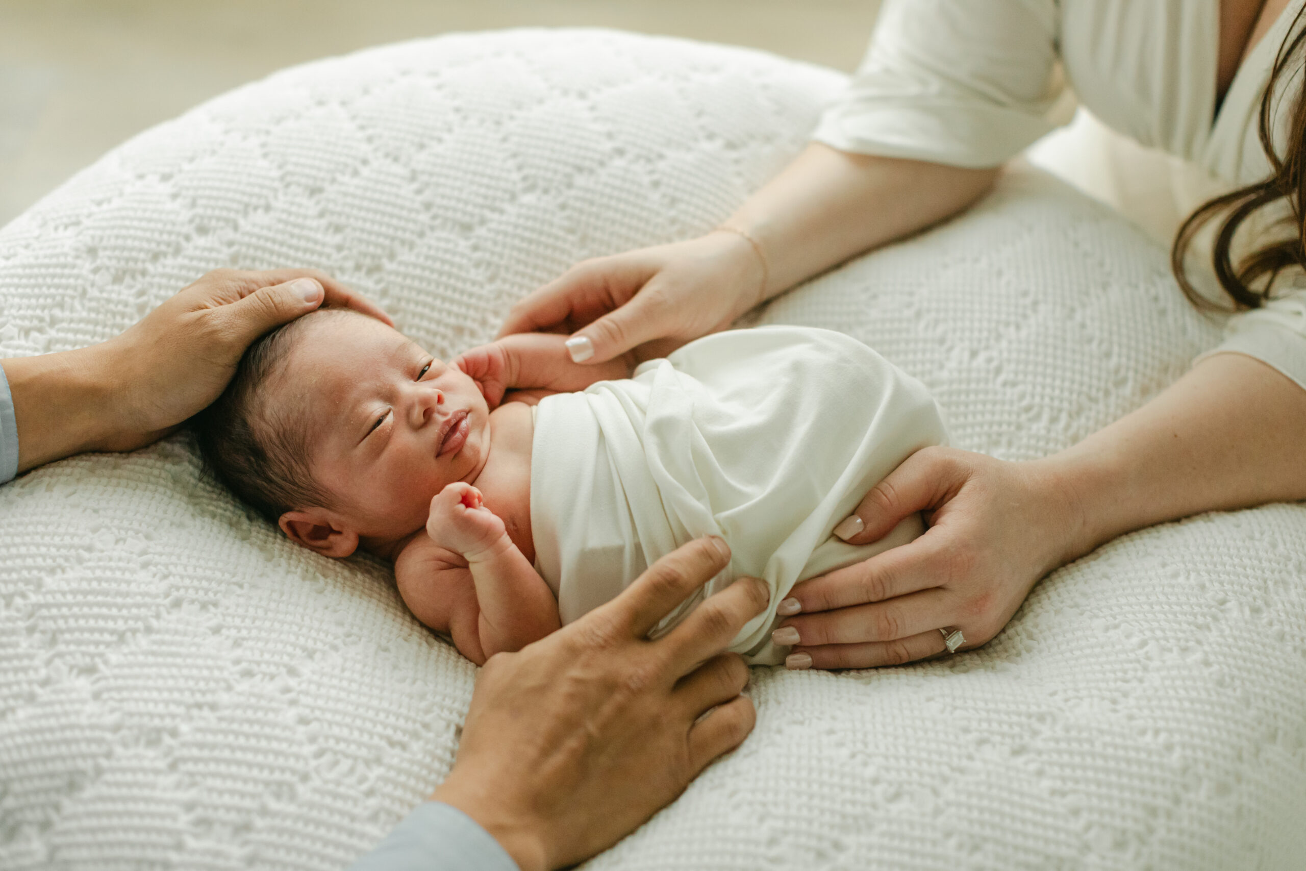 newborn baby in white swaddle blanket, parents hands touching him