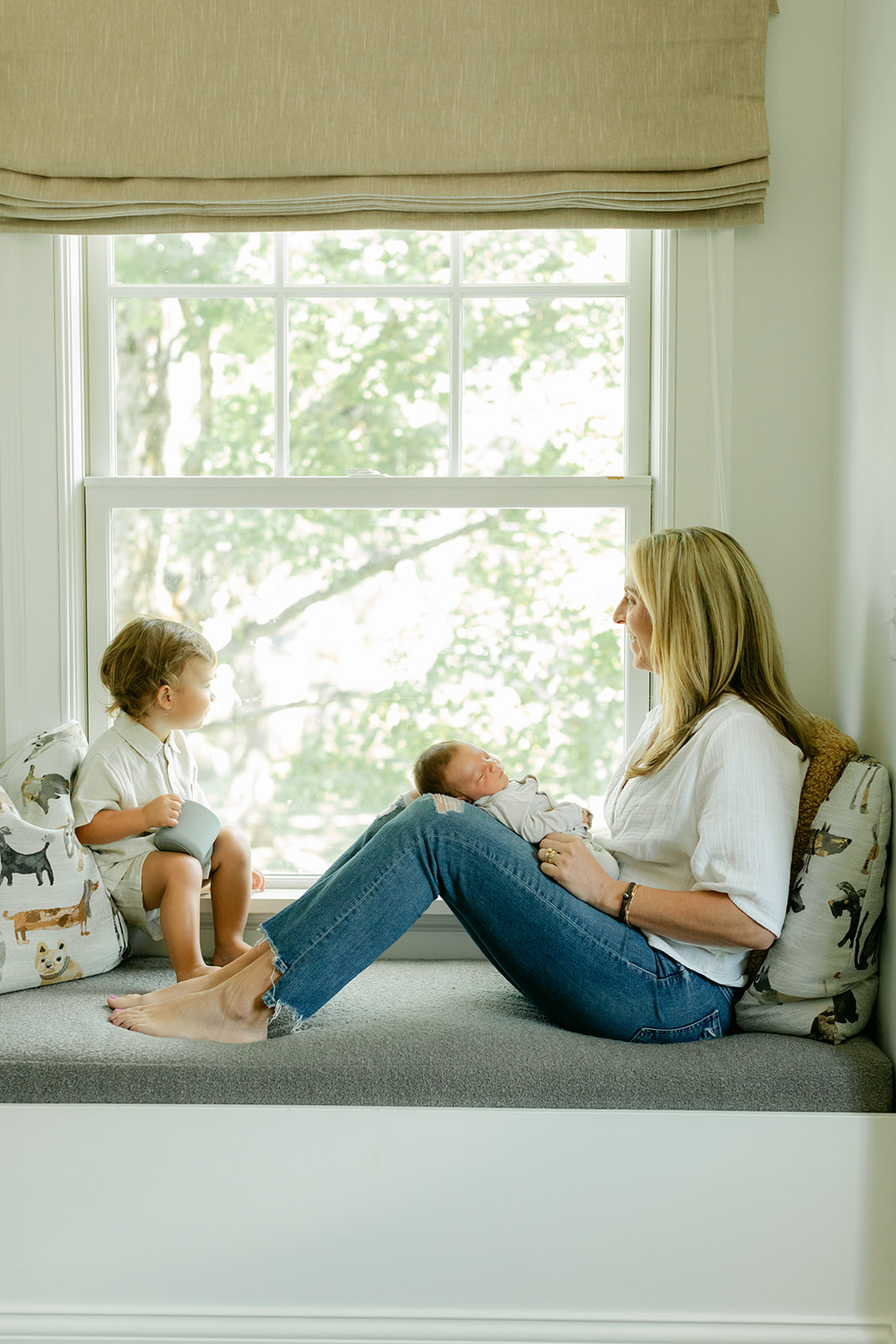 mama in white button up and jeans sitting by window with newborn baby boy and toddler son