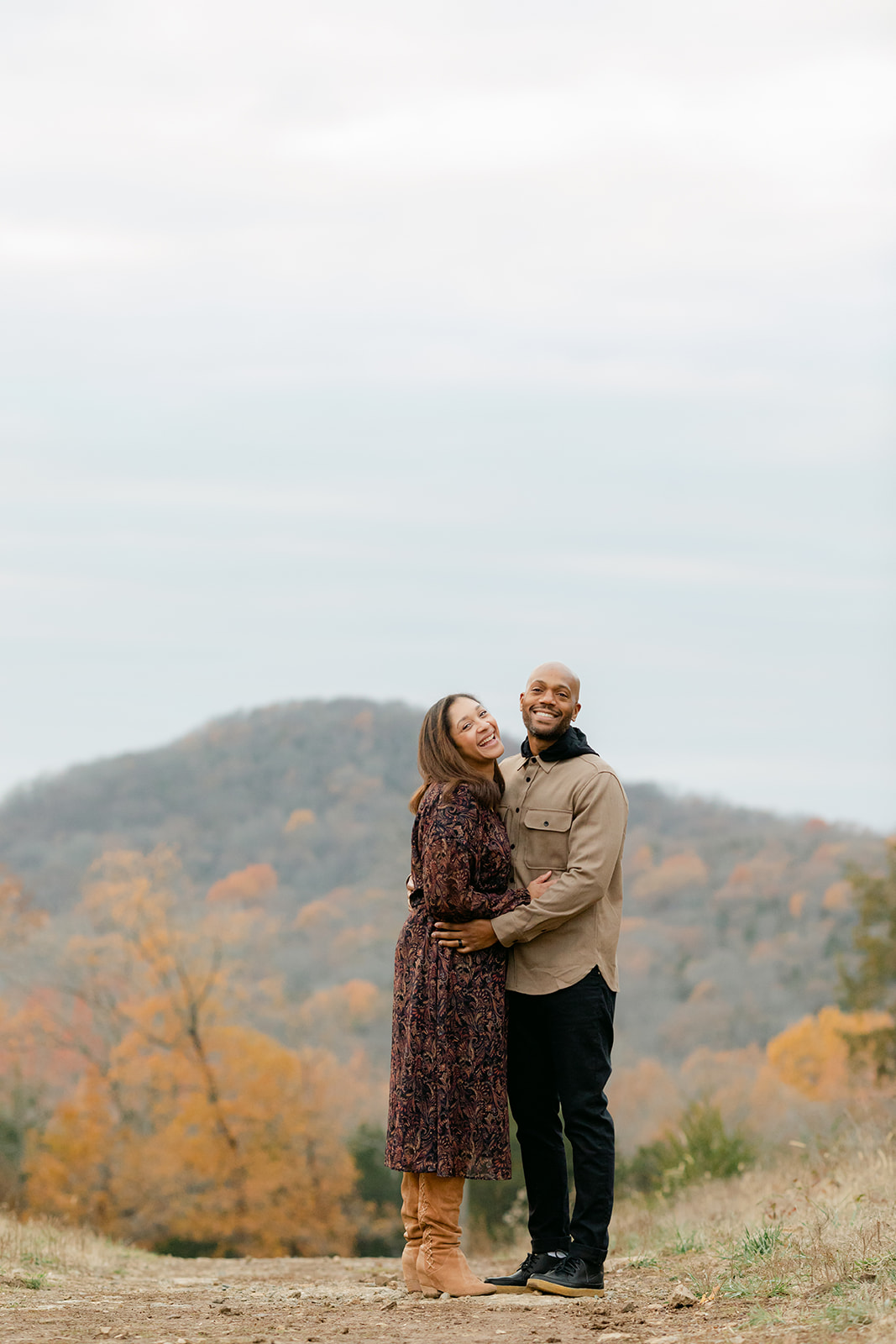 couple stands hugging and laughing at the top of a hill covered with orange fall leaves