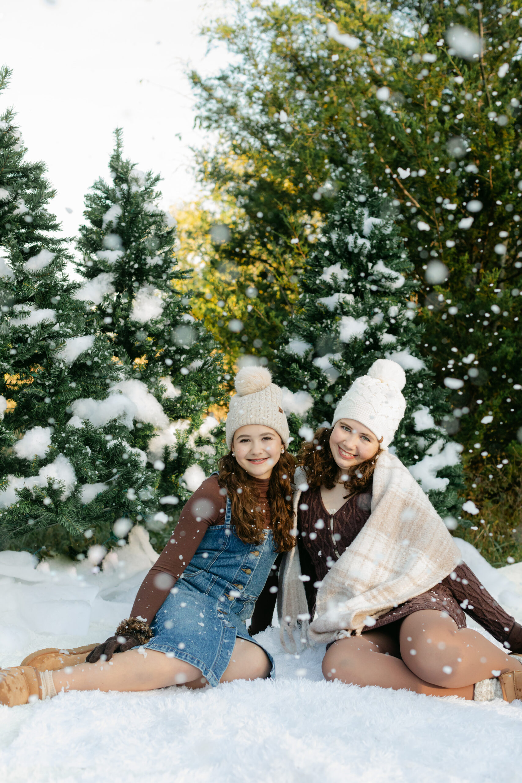 two sisters in cute brown winter outfits wearing beanies laying on the ground surrounded by fake snow for winter christmas photos