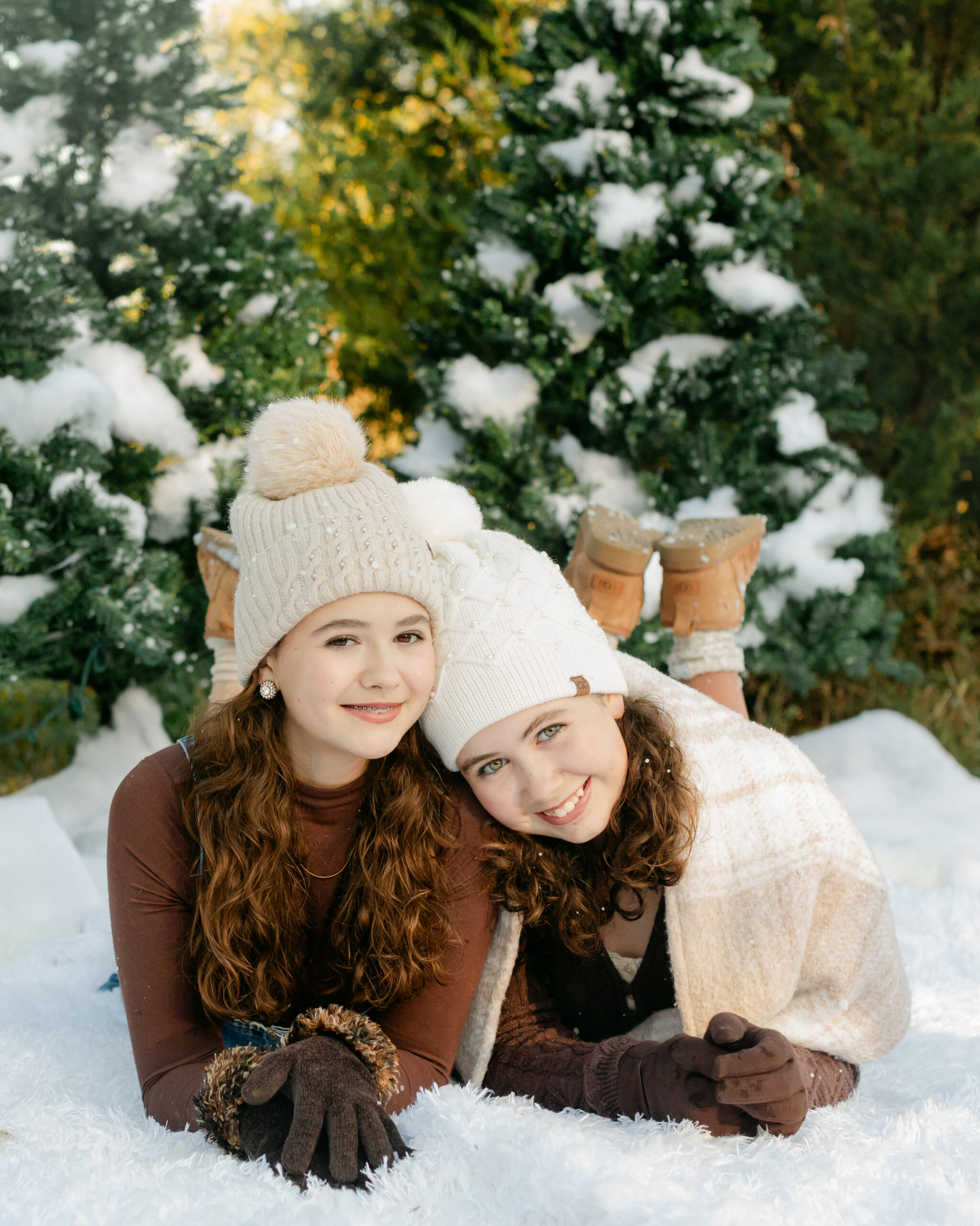 two sisters in cute brown winter outfits wearing beanies laying on the ground surrounded by fake snow for winter christmas photos