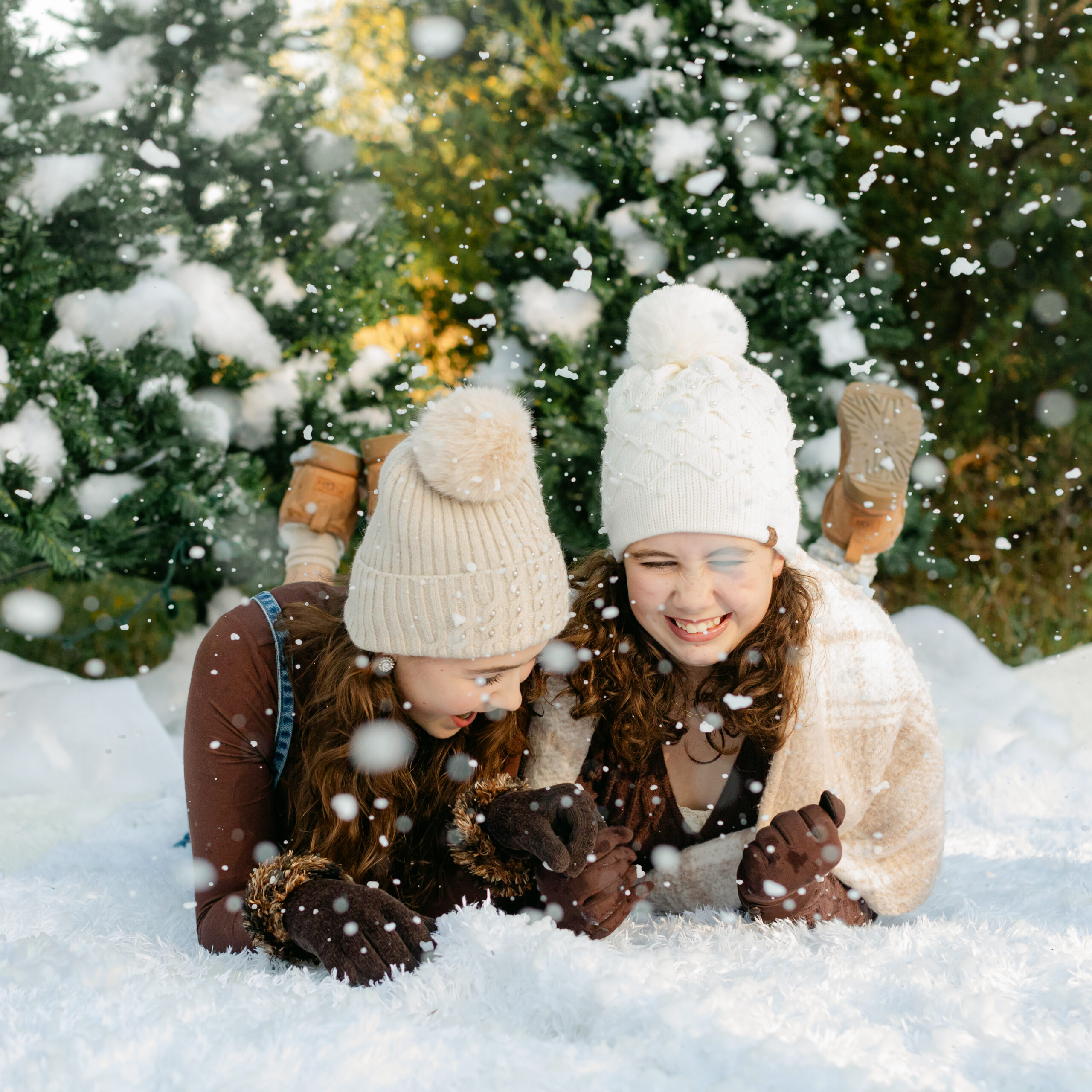 two sisters in cute brown winter outfits wearing beanies laying on the ground surrounded by fake snow for winter christmas photos