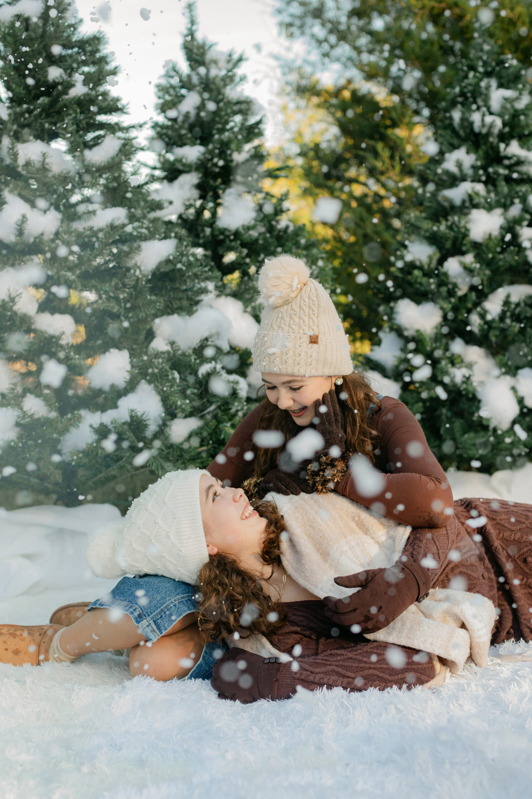 two sisters in cute brown winter outfits wearing beanies laying on the ground surrounded by fake snow for winter christmas photos