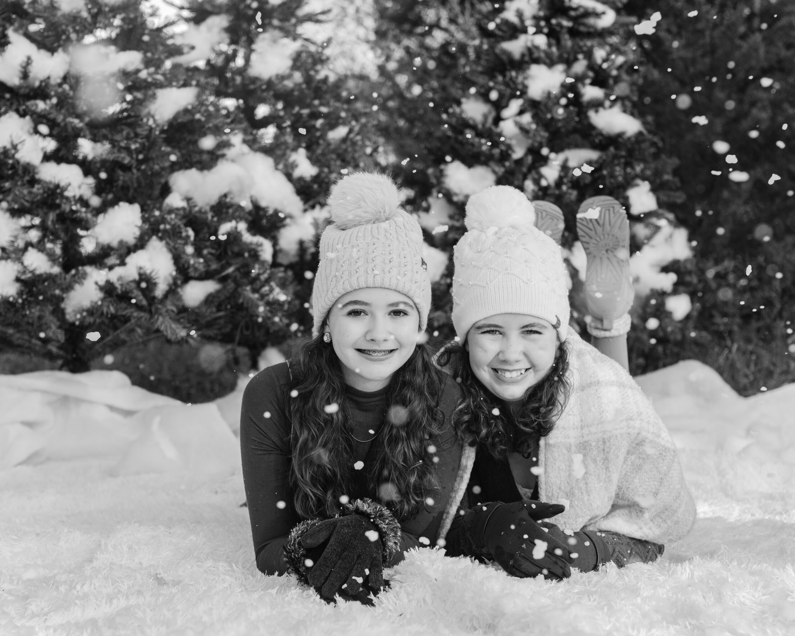 two sisters in cute brown winter outfits wearing beanies laying on the ground surrounded by fake snow for winter christmas photos