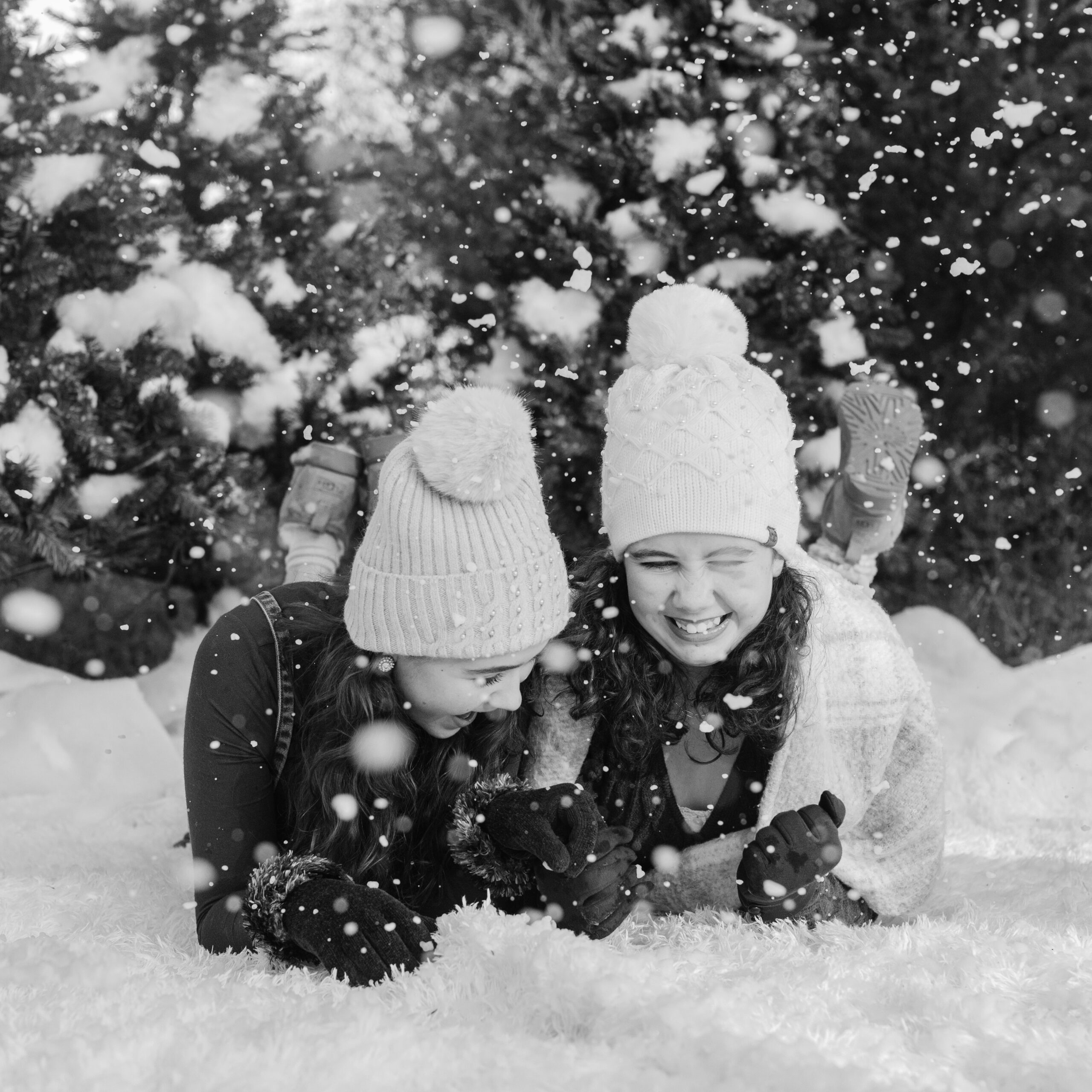 two sisters in cute brown winter outfits wearing beanies laying on the ground surrounded by fake snow for winter christmas photos